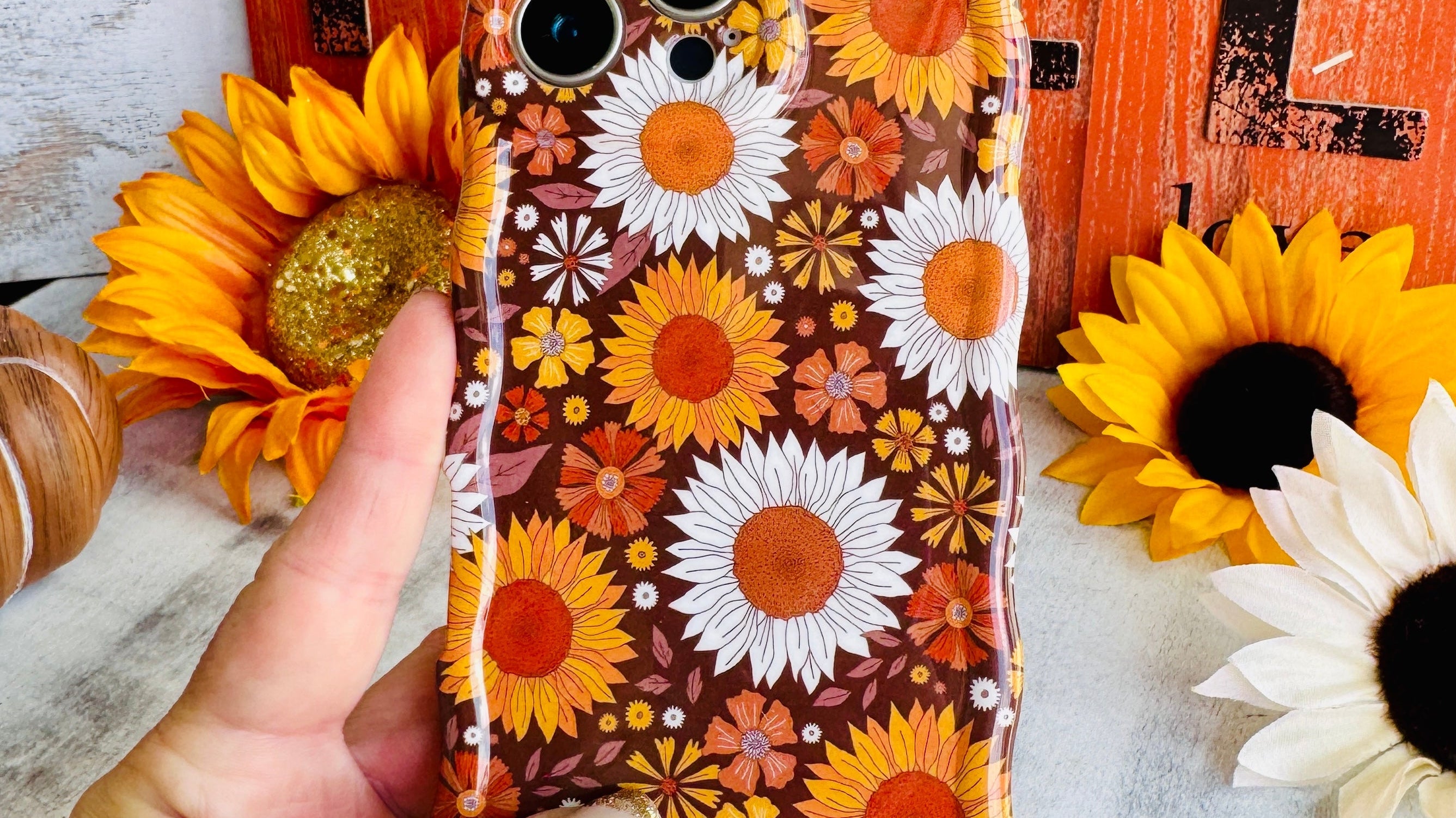 Harvest Sunflowers Wavy Luxe MagSafe iPhone case held against sunflowers and a rust barn door — the Fancy Bands Fall 2026 Collection