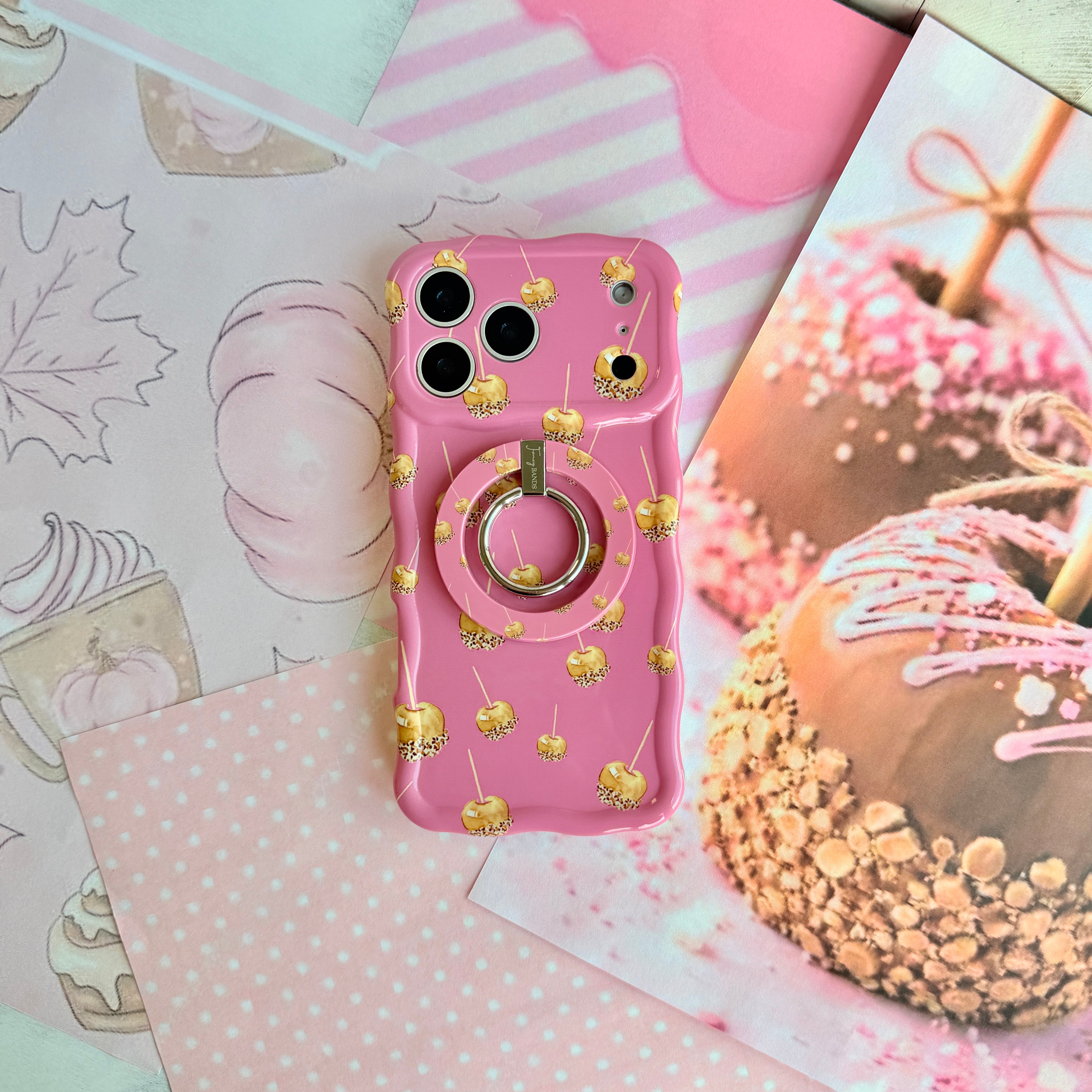 Caramel Coated Cutie Wavy Luxe iPhone case shown with the matching Caramel Coated Cutie MagSafe grip ring attached, styled beside a real caramel apple rolled in chopped nuts.