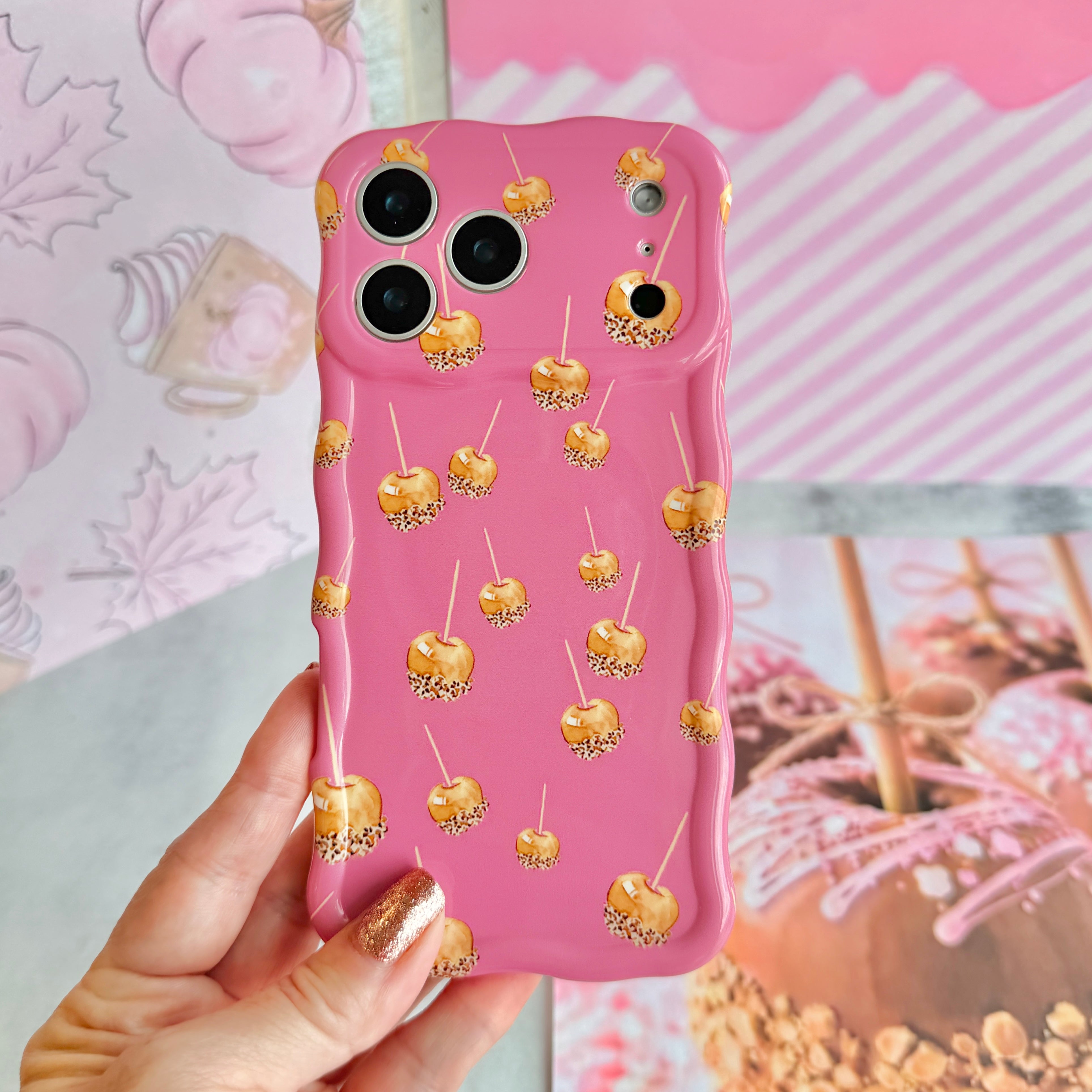 Hand holding the Caramel Coated Cutie Wavy Luxe MagSafe iPhone case, showing the rose-pink base scattered with golden caramel apples against a pink fall mood board backdrop.