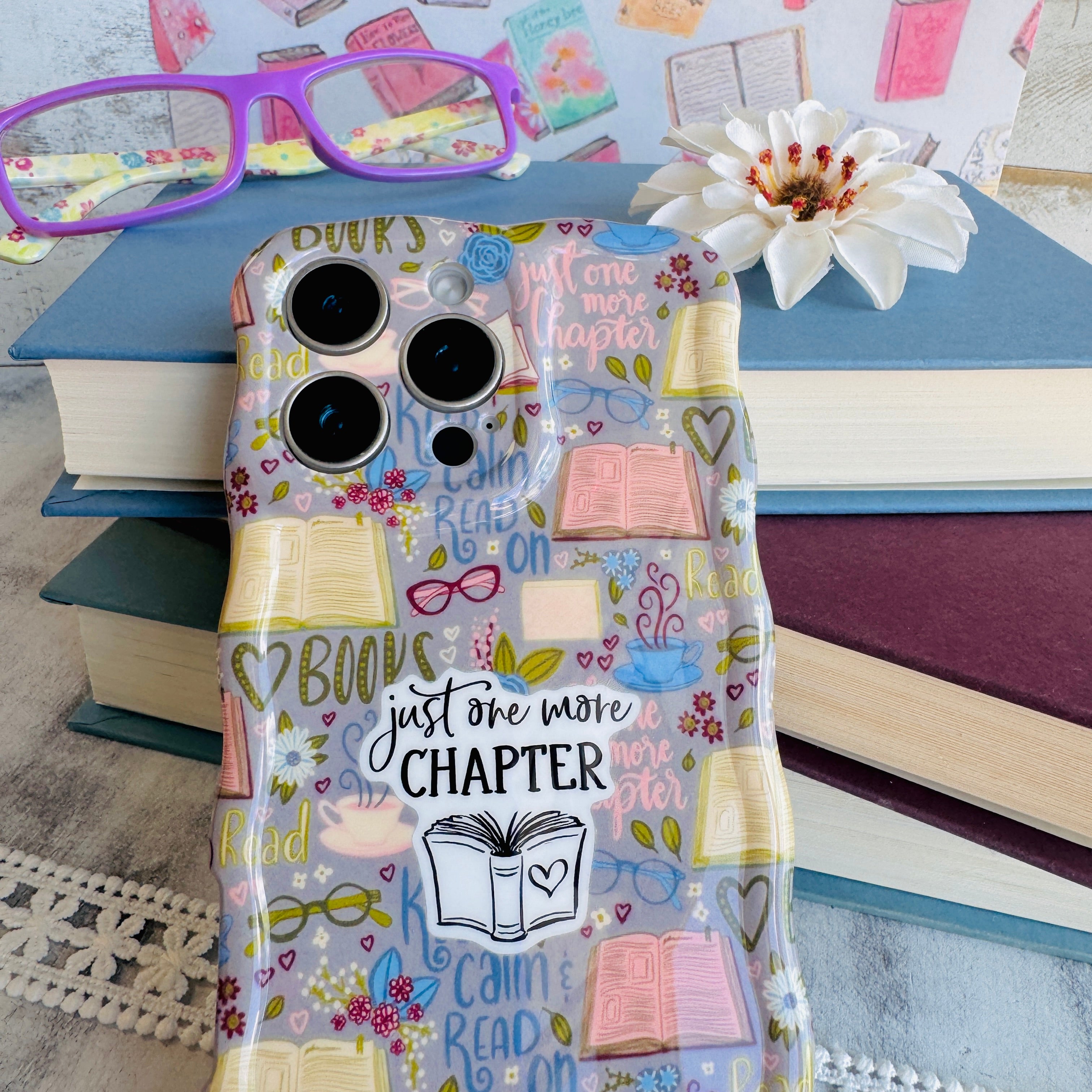 Phone case with book-themed design on a stack of books with glasses and flowers.