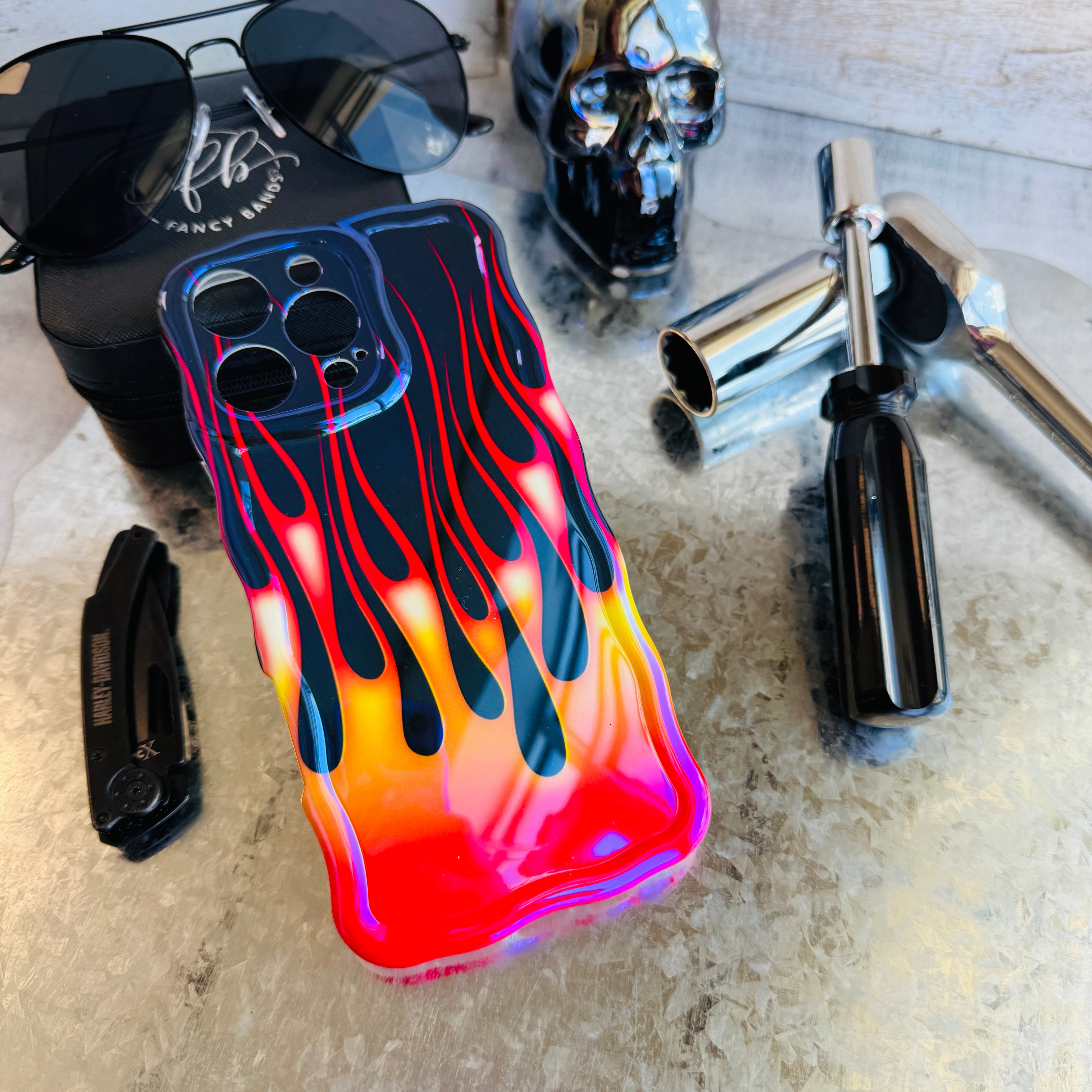 Phone case with flame design on a surface with sunglasses, skull, and bottle.