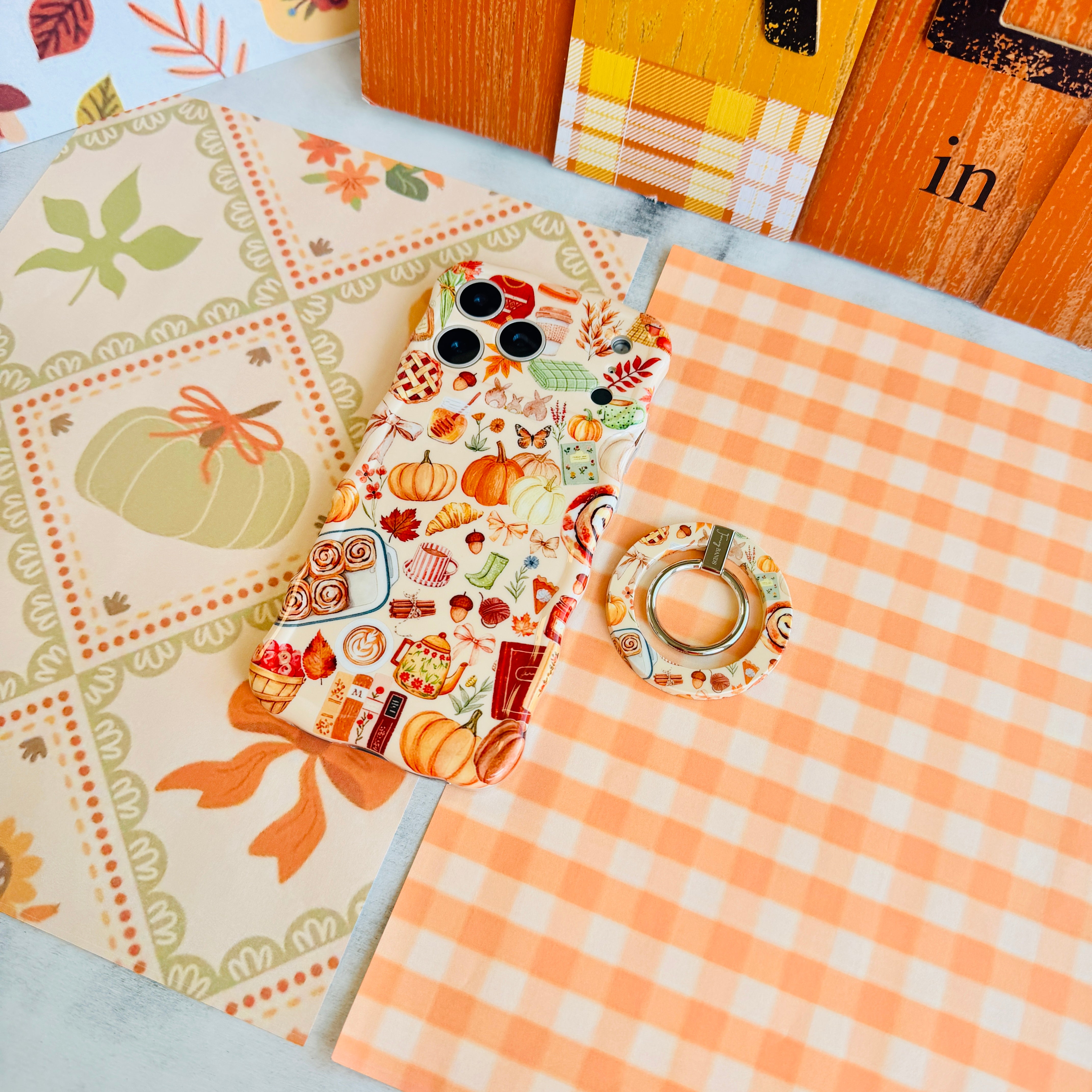 All The Fall Things MagSafe grip ring shown beside the matching All The Fall Things iPhone case on orange gingham and cream fall paper.