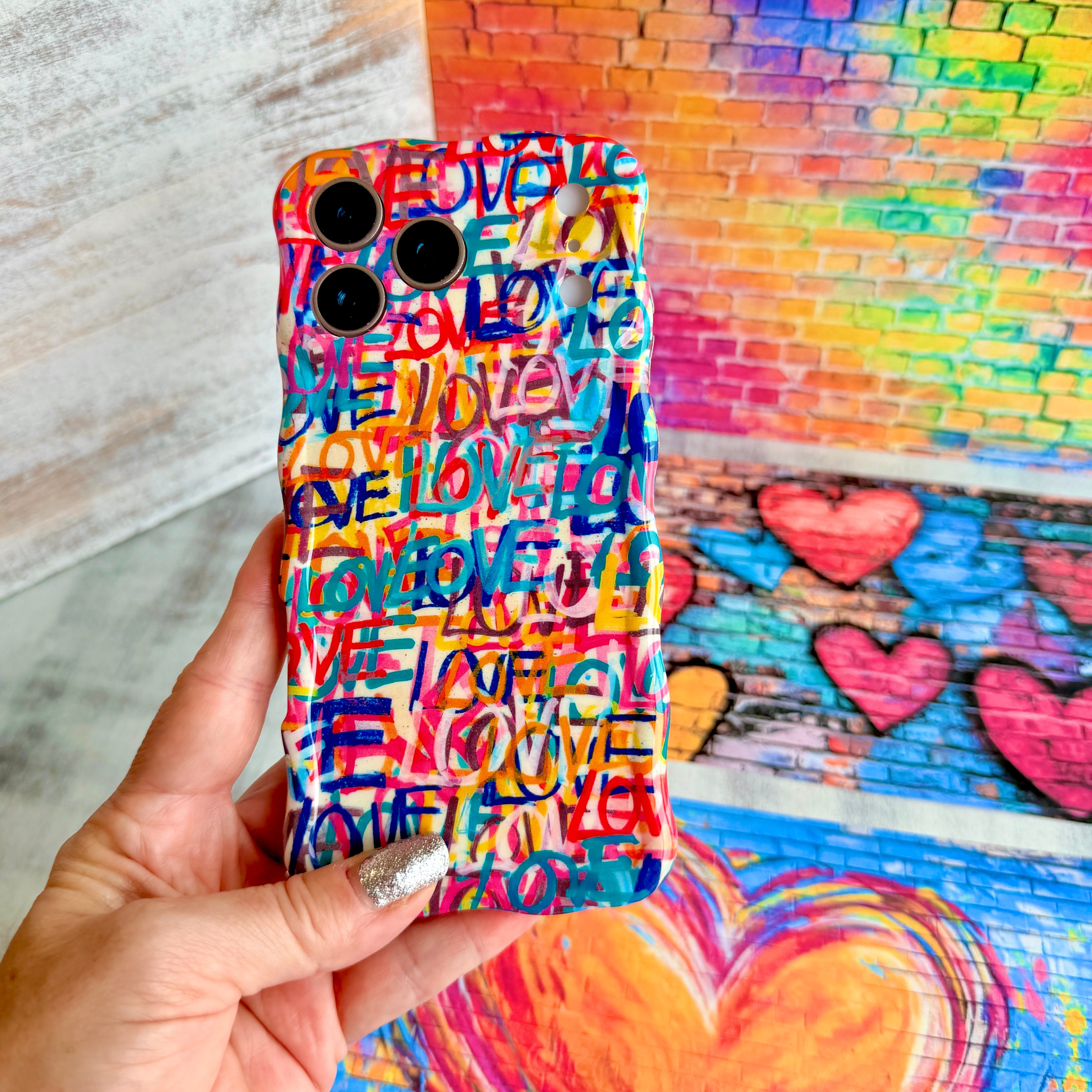 Hand holding the Love Riot Wavy Luxe MagSafe iPhone case with graffiti lettering and painted hearts on a brick wall background