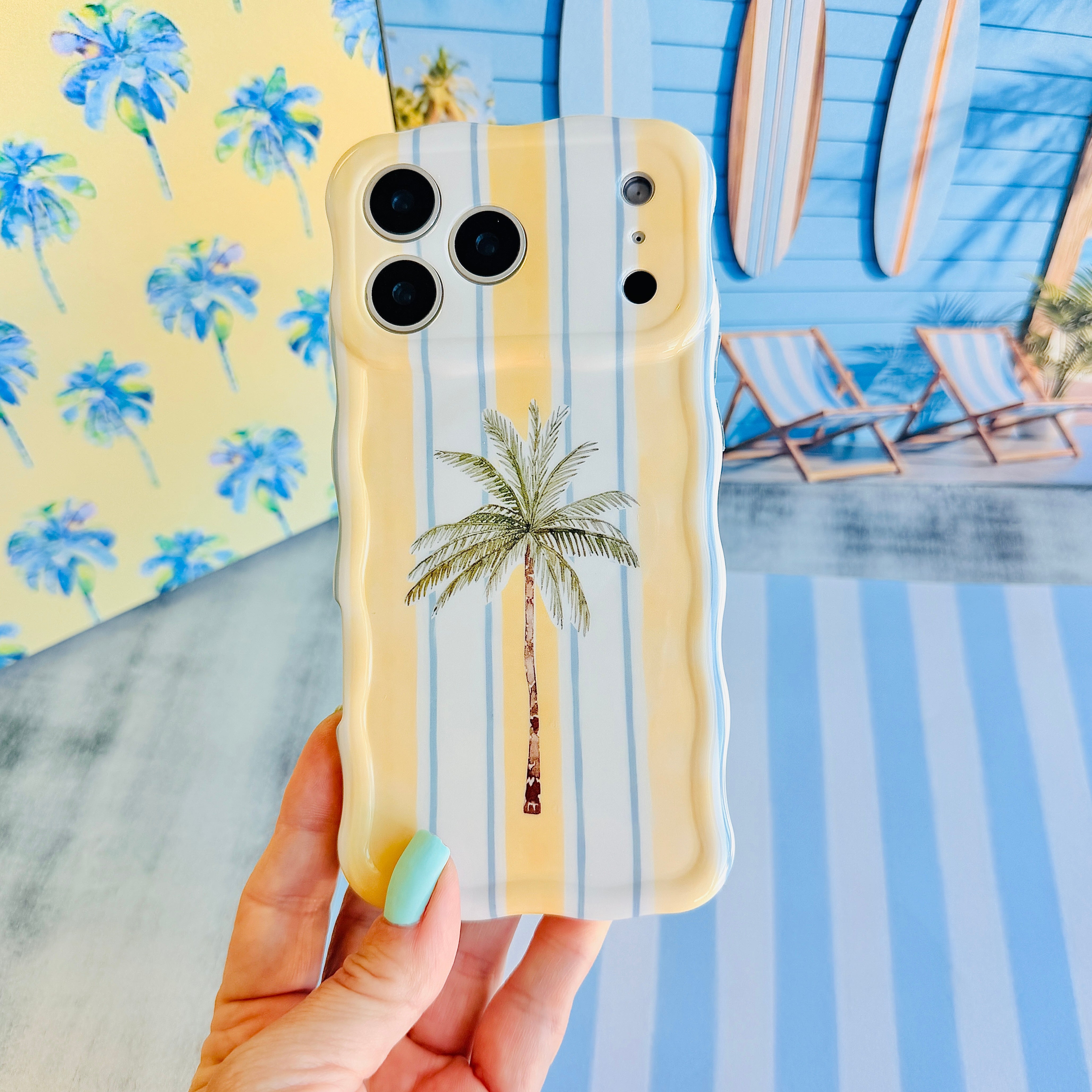 Hand holding the Palm Shore Club Wavy Luxe MagSafe iPhone case with a hand-drawn palm on butter-yellow and sky-blue cabana stripes