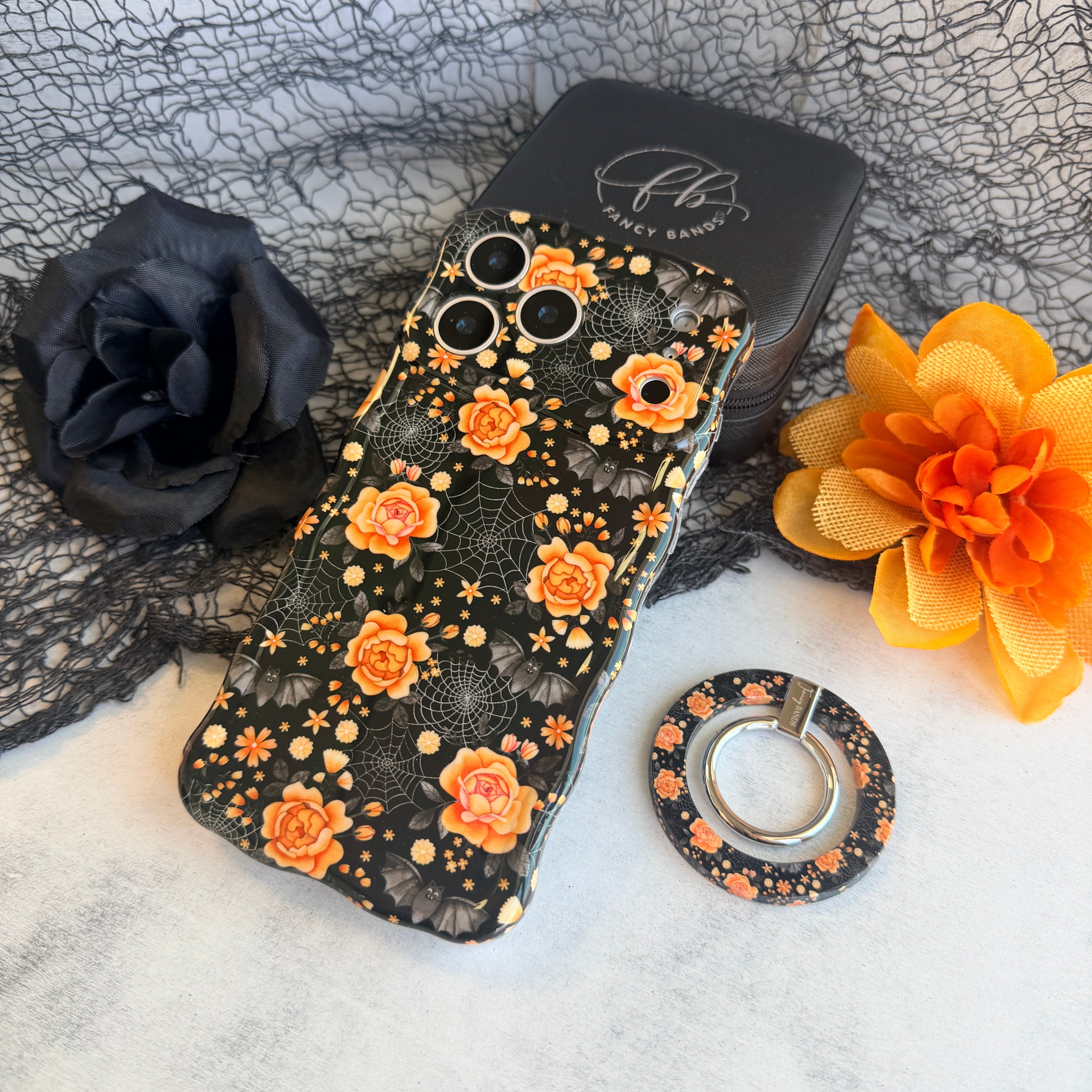 Haunted Blossoms MagSafe grip ring lying beside the matching black gothic floral Wavy Luxe case on black spiderweb mesh.