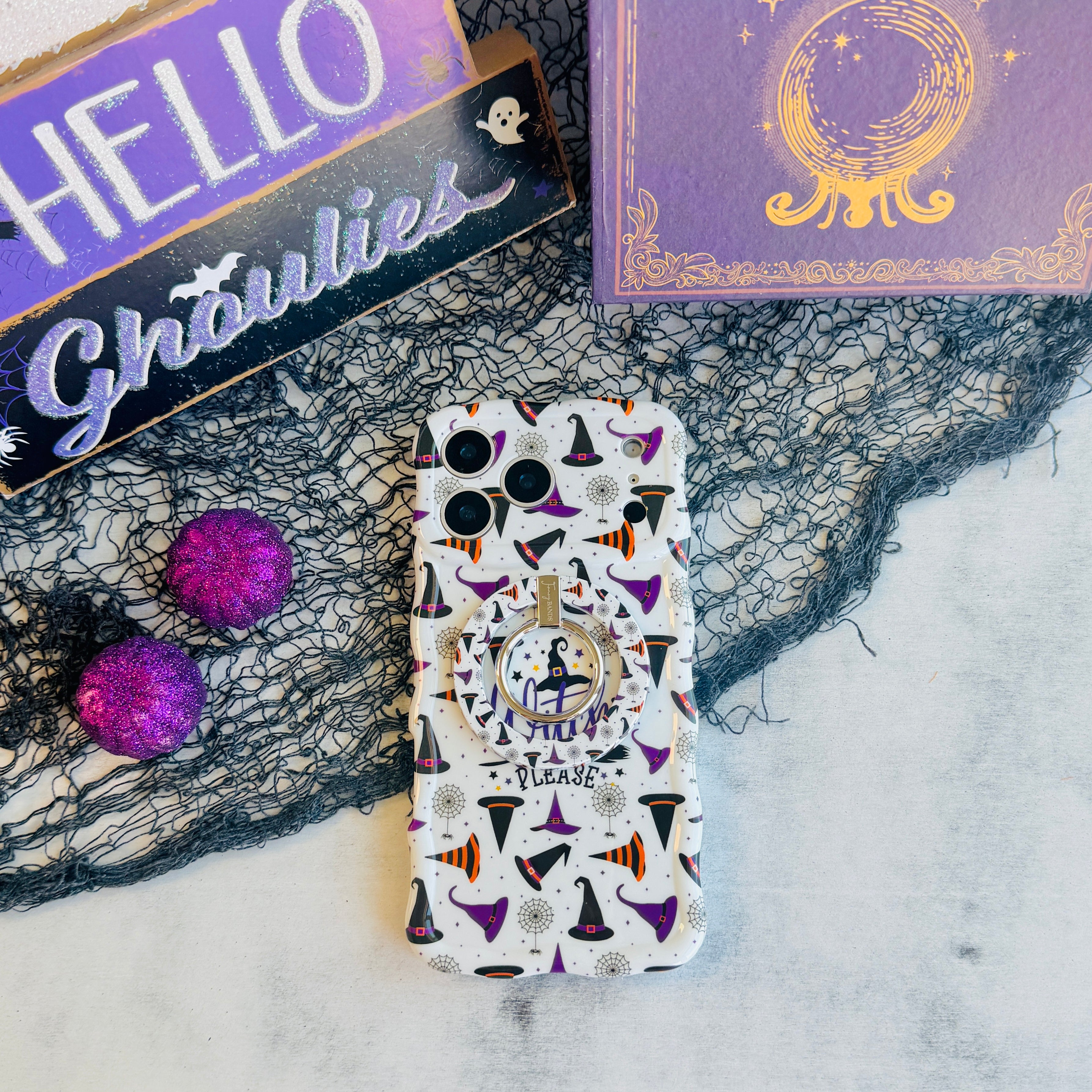 Witch Please MagSafe grip ring attached to the matching cream witch hat Wavy Luxe case, shown with a HELLO Ghoulies sign and Book of Spells backdrop.