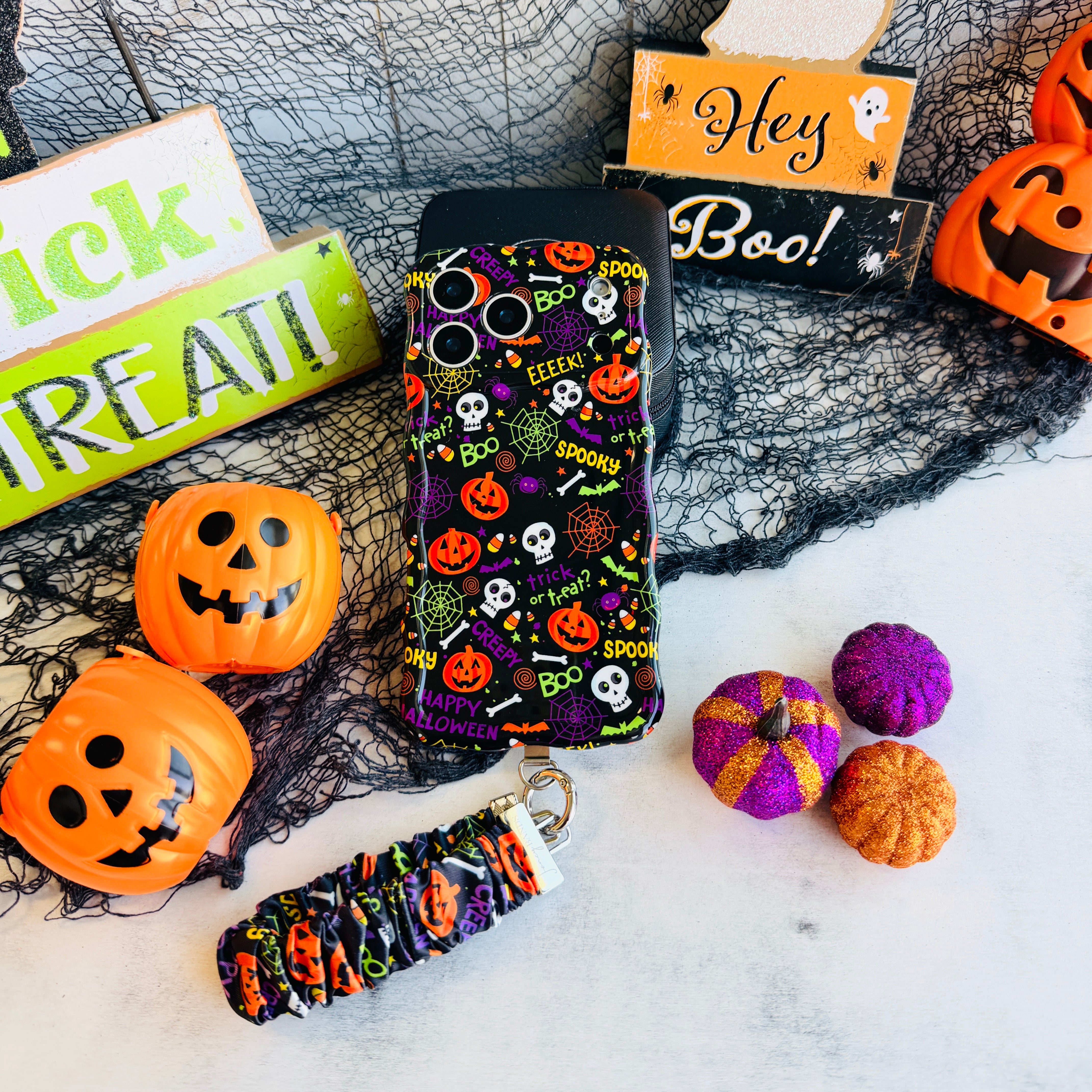 Spooky scrunchie phone wrist strap and matching Wavy Luxe case styled together with a Hey Boo sign and orange jack-o-lantern pails.