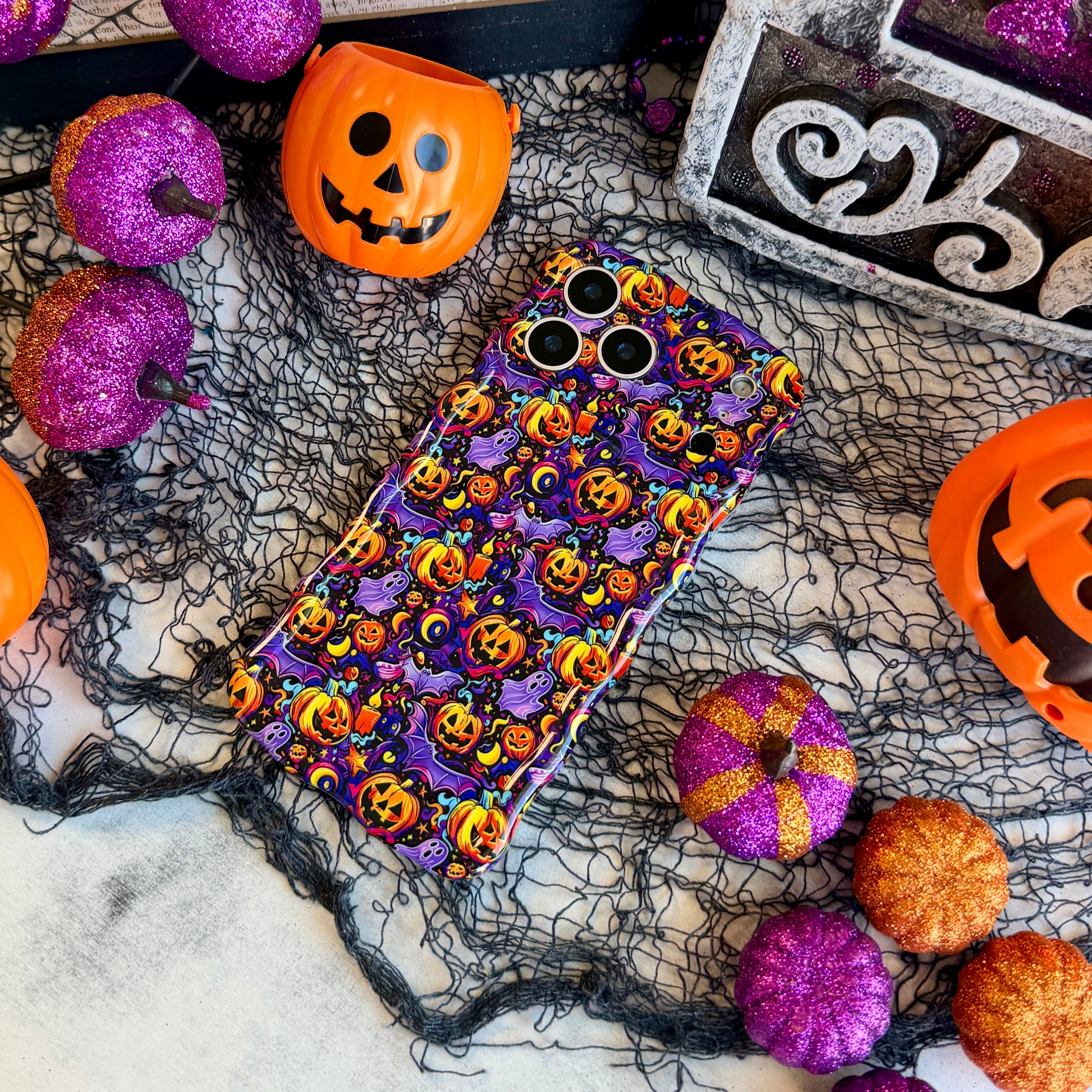 Jack-O-Lantern Jive Wavy Luxe MagSafe iPhone case laid flat on black spiderweb mesh with orange jack-o-lantern pails and glitter pumpkins.