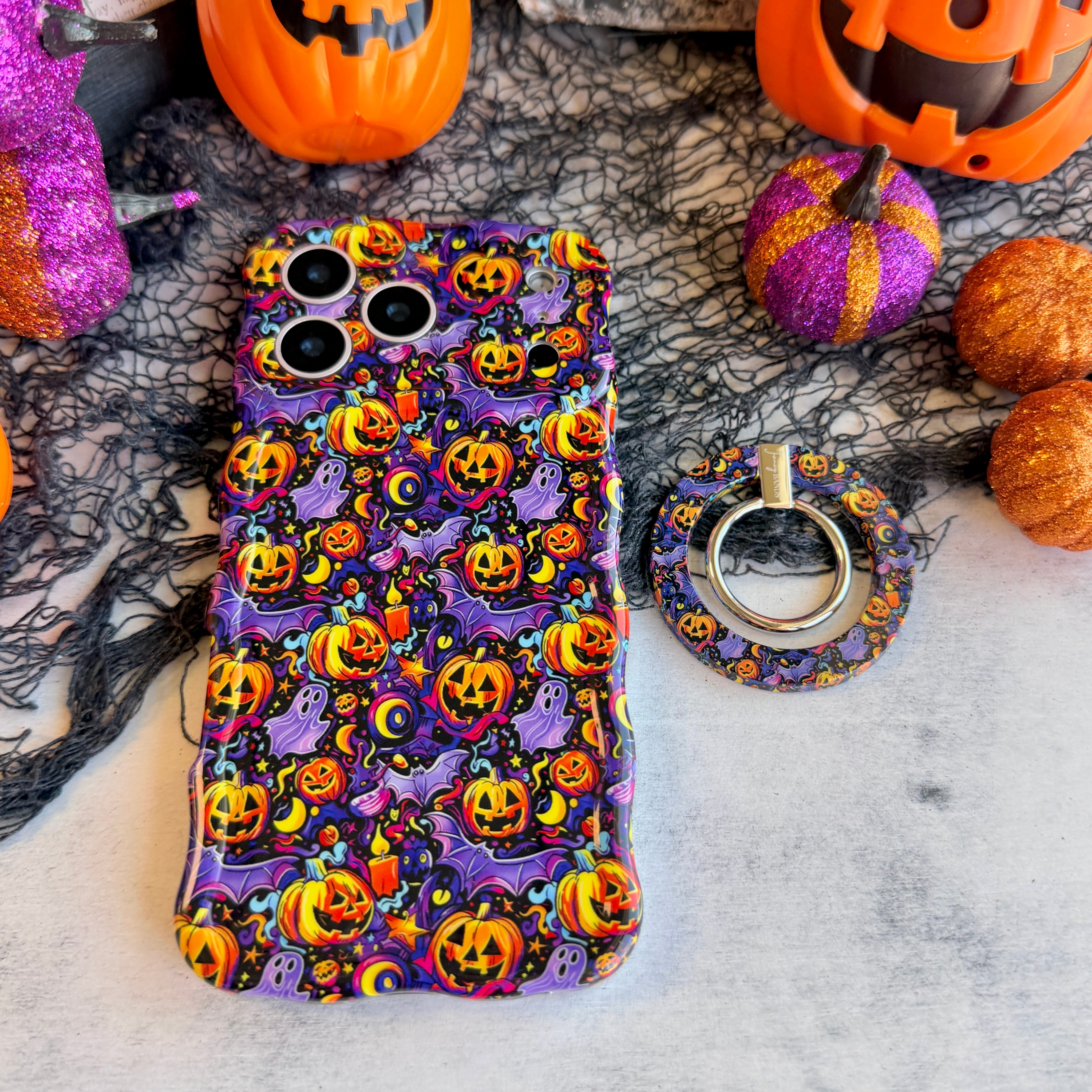 Jack-O-Lantern Jive MagSafe grip ring lying beside the matching Wavy Luxe case on marble, showing the violet neon pumpkin print on both.
