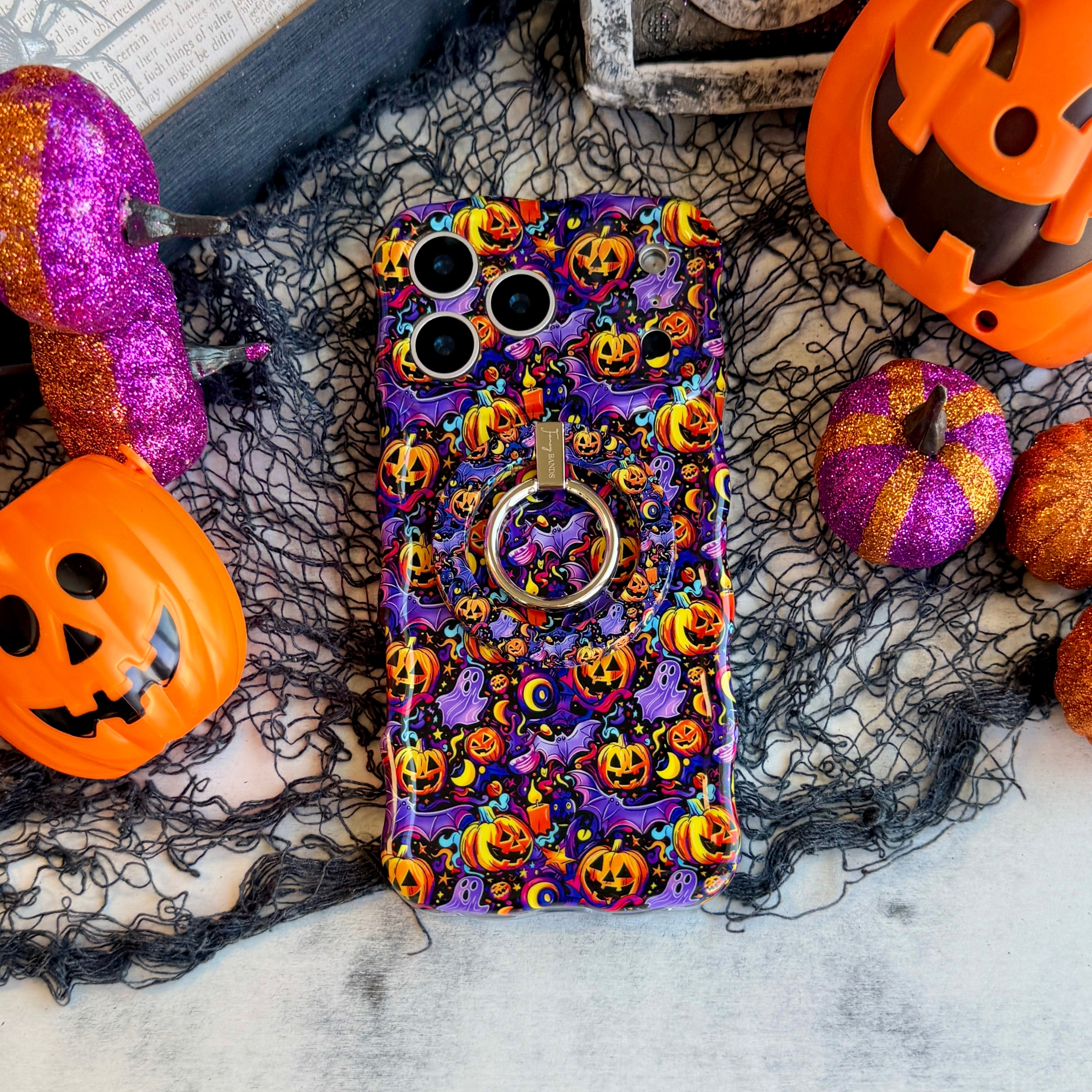 Jack-O-Lantern Jive Wavy Luxe MagSafe iPhone case with the matching MagSafe grip ring attached, styled with orange pails and magenta glitter pumpkins.