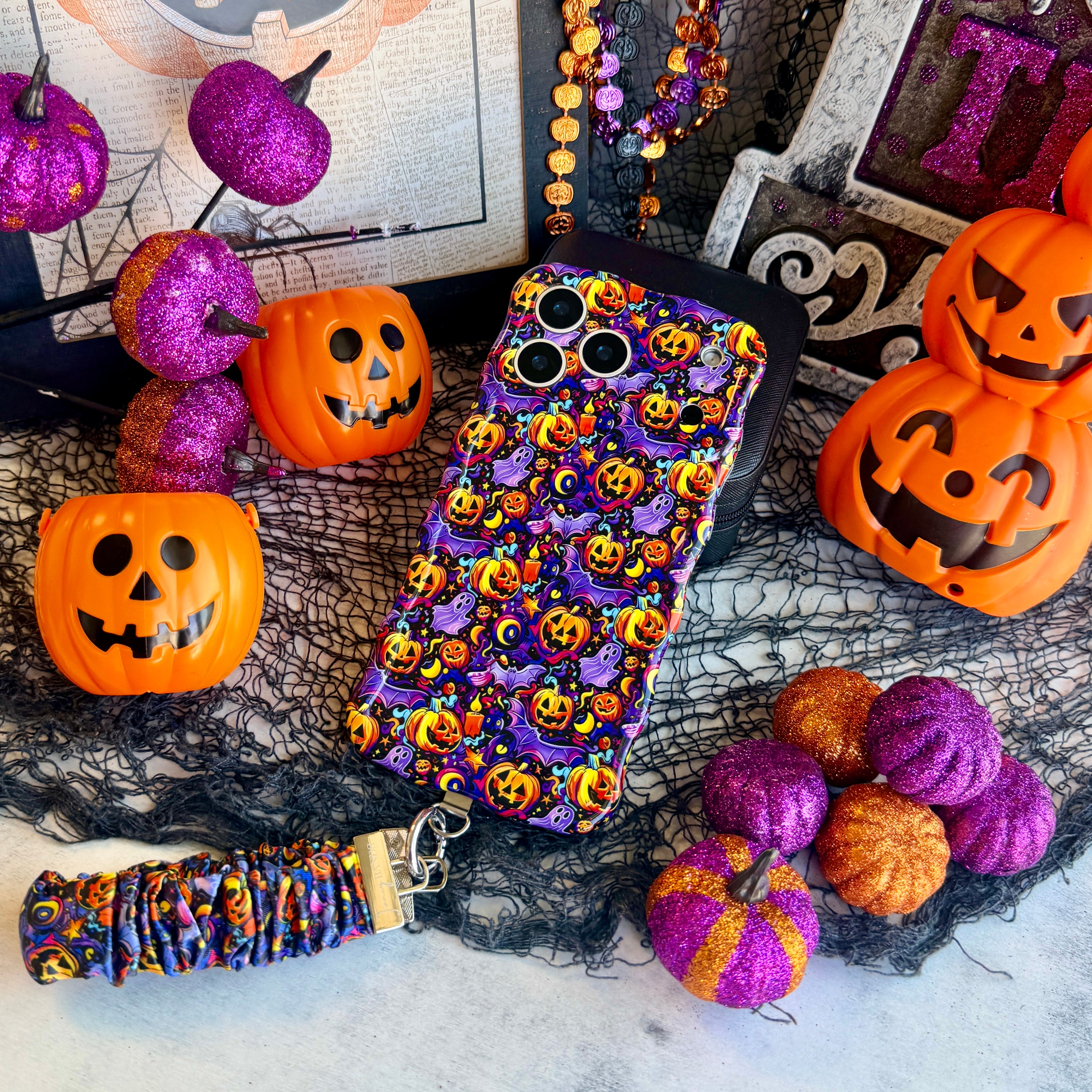 Jack-O-Lantern Jive scrunchie phone wrist strap and matching Wavy Luxe case styled together on a Halloween set with pumpkins and a beaded garland.