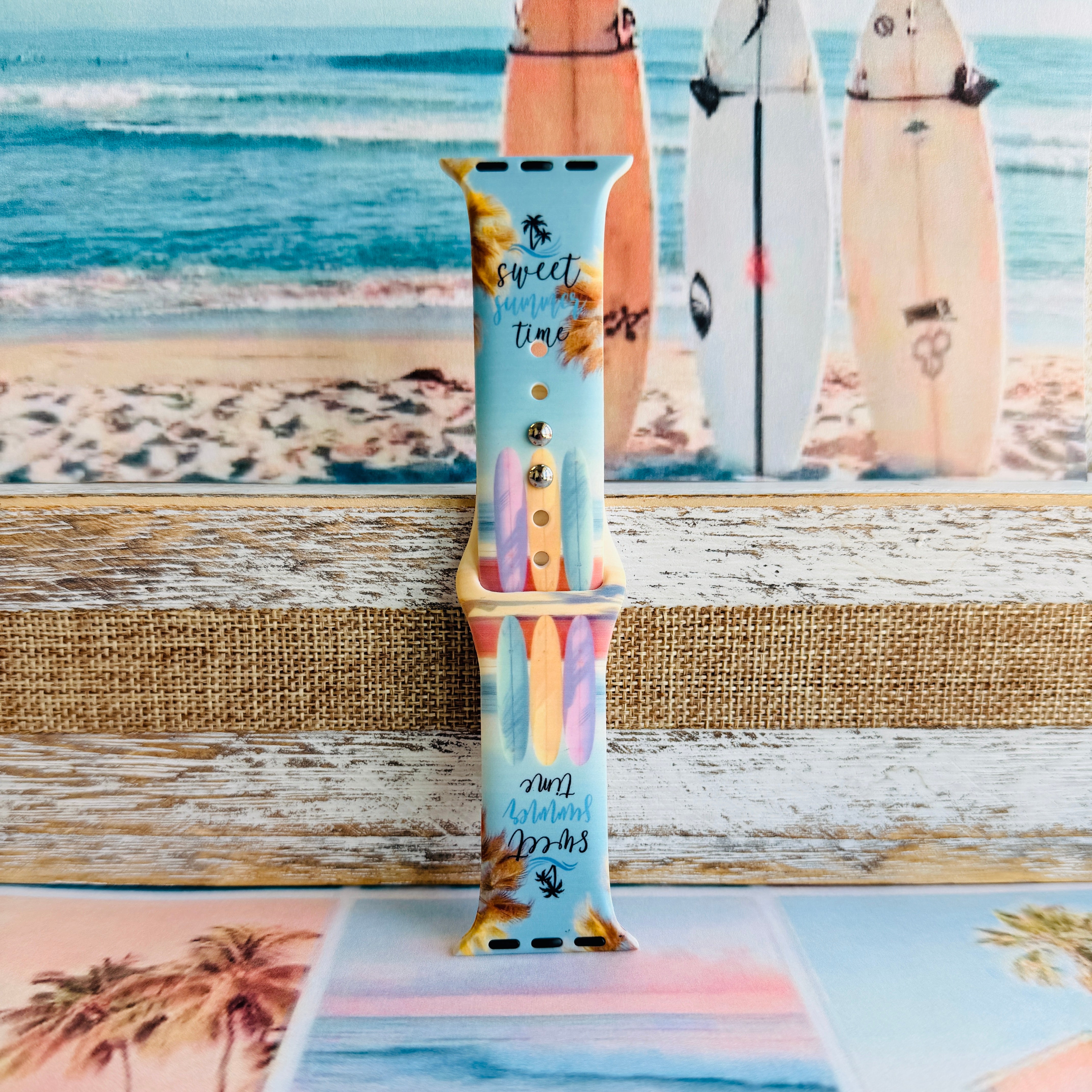 Sweet Summer silicone Apple Watch band with pastel pink, yellow, and blue surfboards, palm trees, and "sweet summer time" script on a soft watercolor beach scene, shown displayed and worn on Apple Watch against a coastal surfboard backdrop.