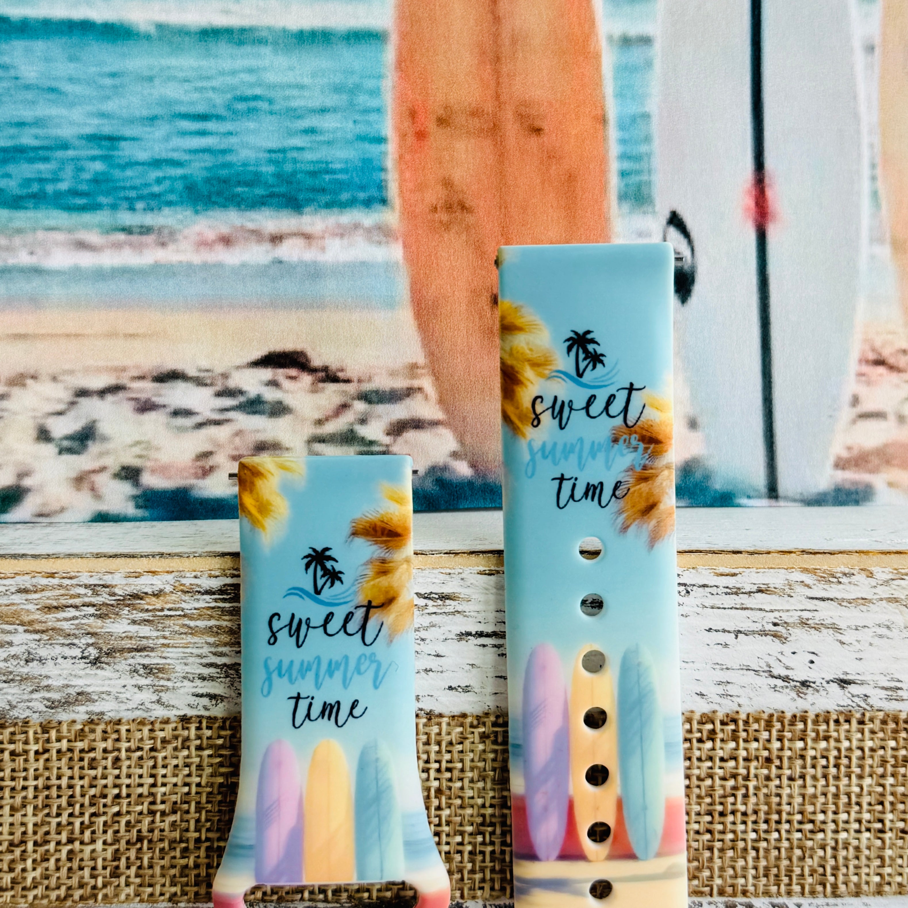 Sweet Summer silicone Samsung Galaxy Watch band with pastel pink, yellow, and blue surfboards, palm trees, and "sweet summer time" script on a soft watercolor beach scene, shown displayed and worn against a coastal surfboard backdrop.