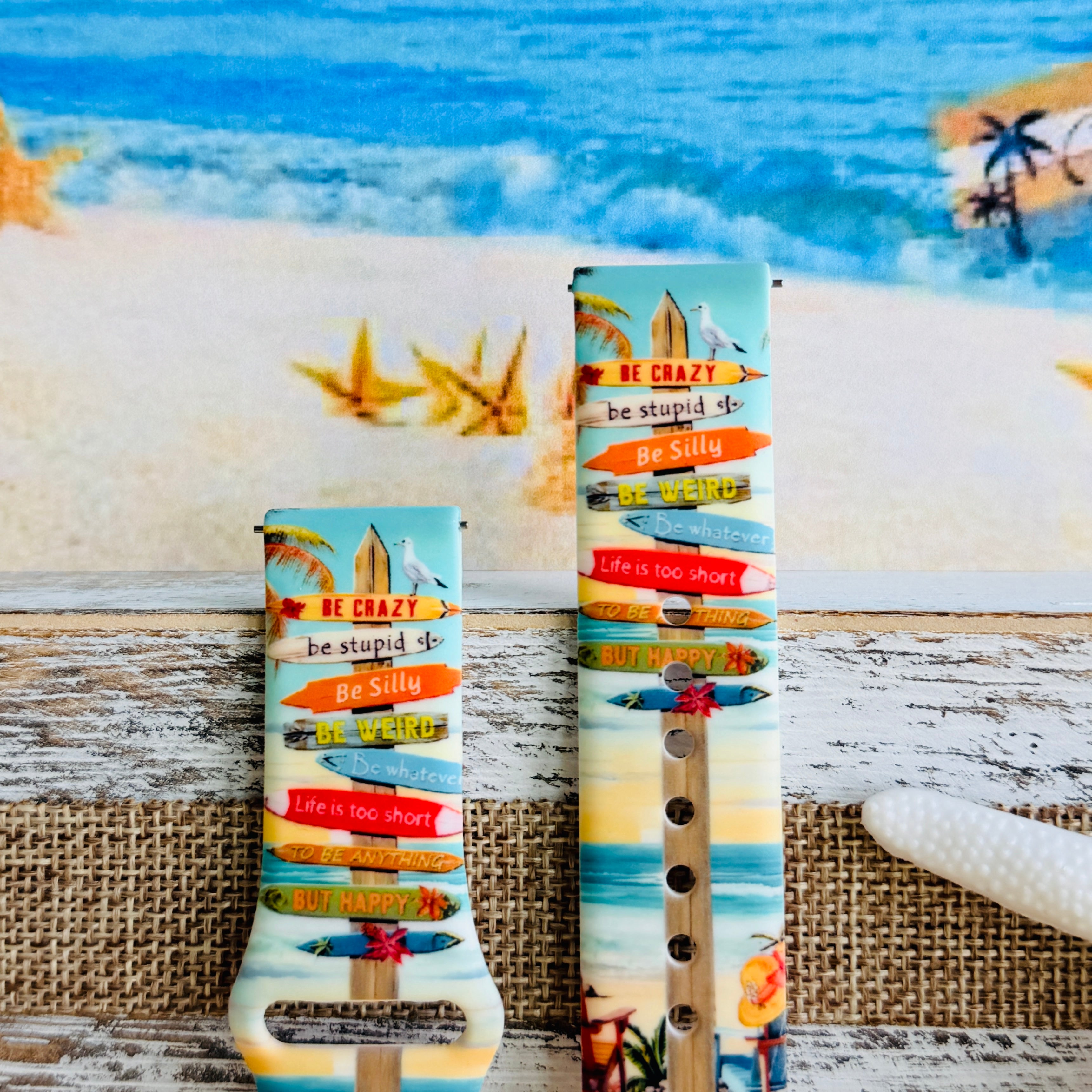 Seaside State of Mind silicone Samsung Watch band with colorful surfboard signpost on a beach scene and "Be Crazy, Be Silly, Be Weird—life is too short to be anything but happy" message, shown displayed and worn on Apple Watch against a beach backdrop.