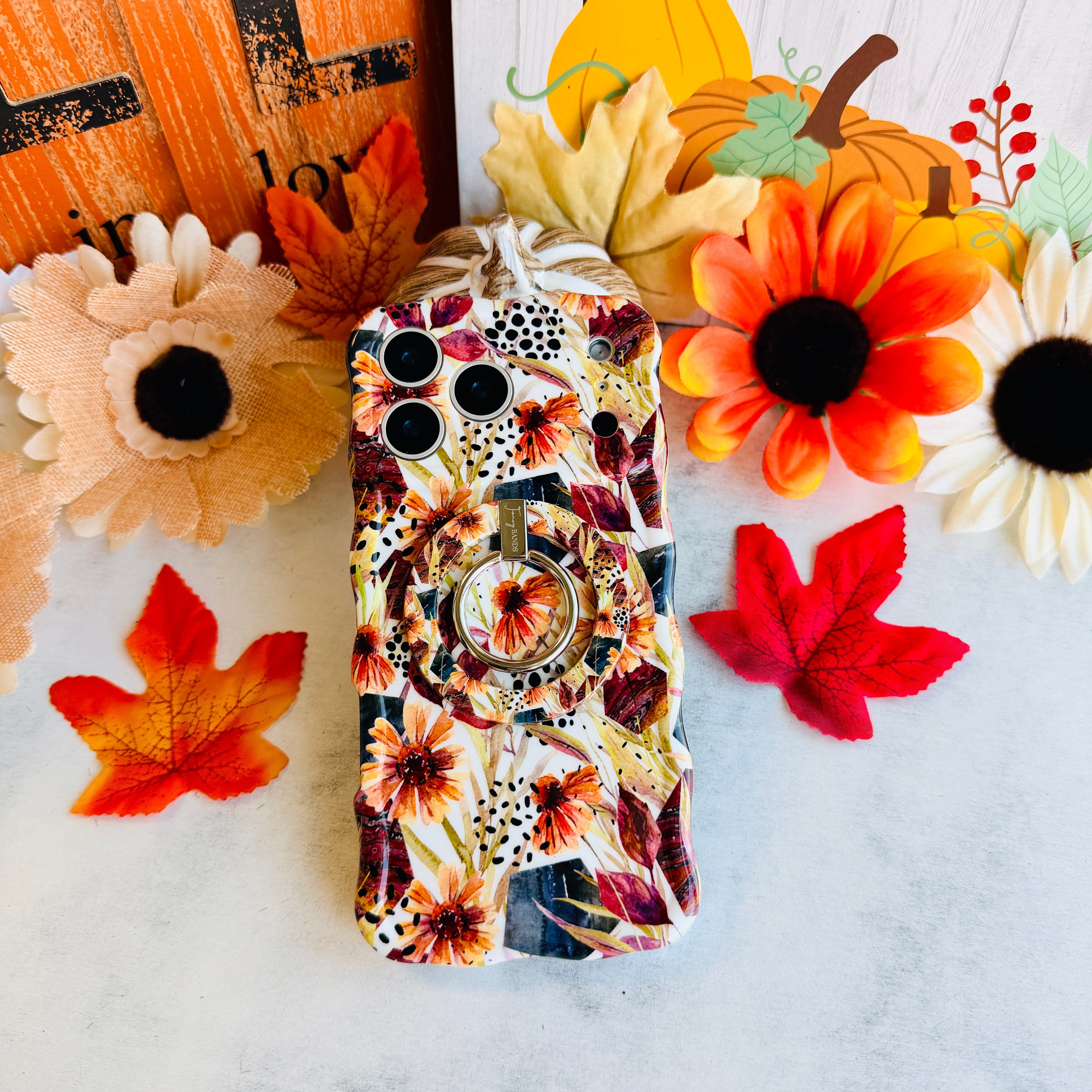 Sienna Petals Wavy Luxe iPhone case with the matching MagSafe grip ring attached, styled with sunflowers and maple leaves on white marble.