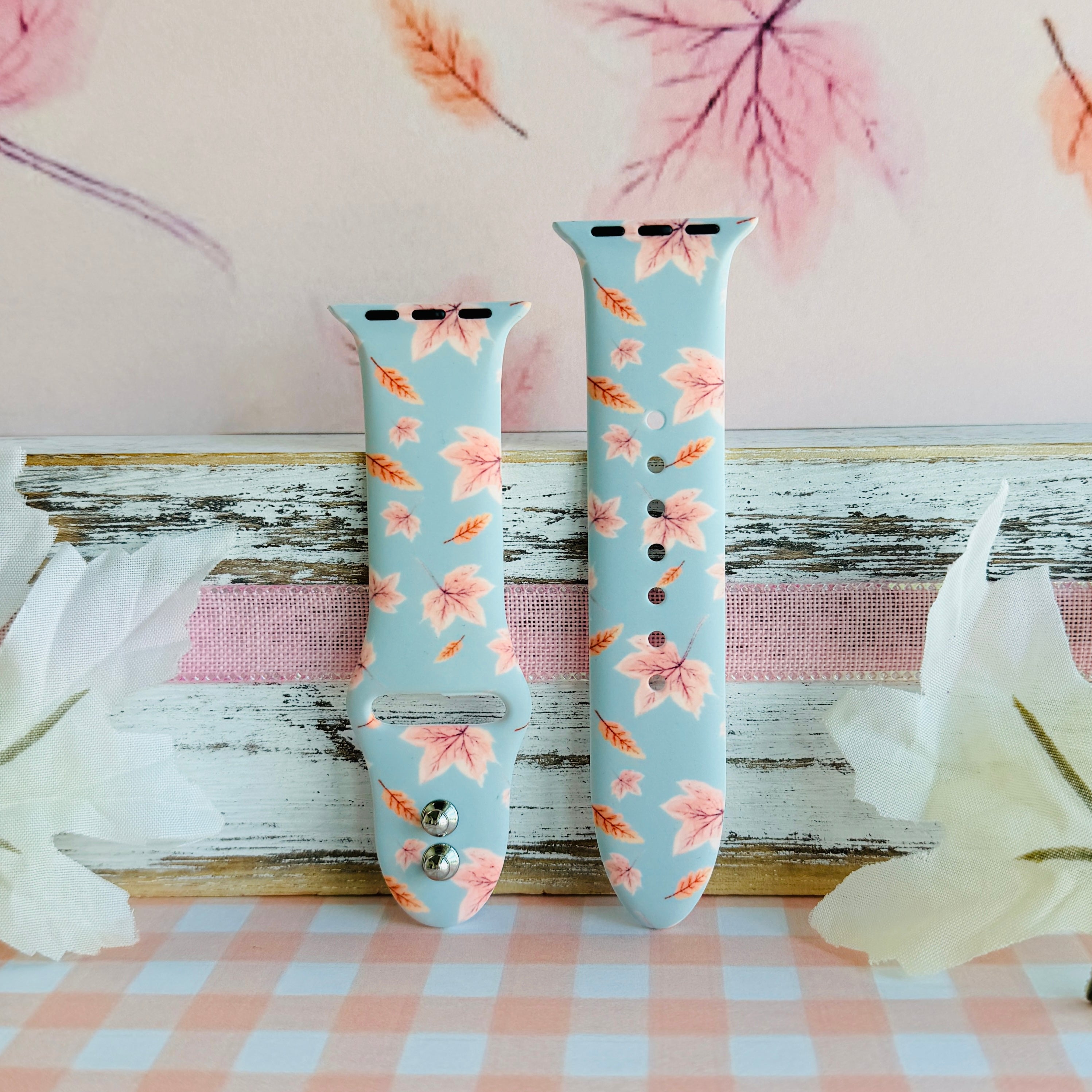 Autumn Breeze Apple Watch band in both size options on a weathered wood ledge with pale silk leaves and a pink gingham cloth.