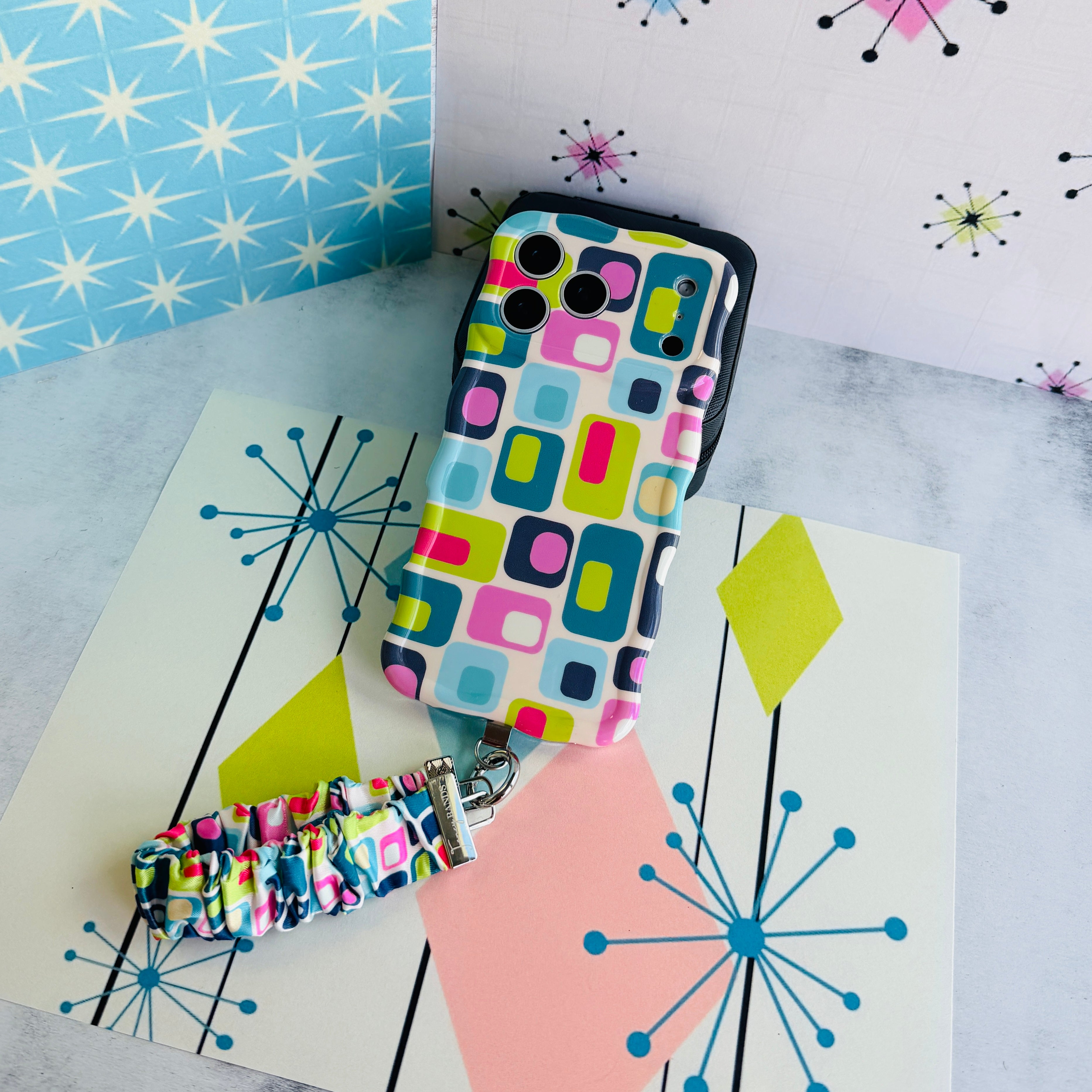 Mod Mood Scrunchie Phone Wrist Strap and matching Wavy Luxe phone case laid out together on a mid-century starburst print backdrop.