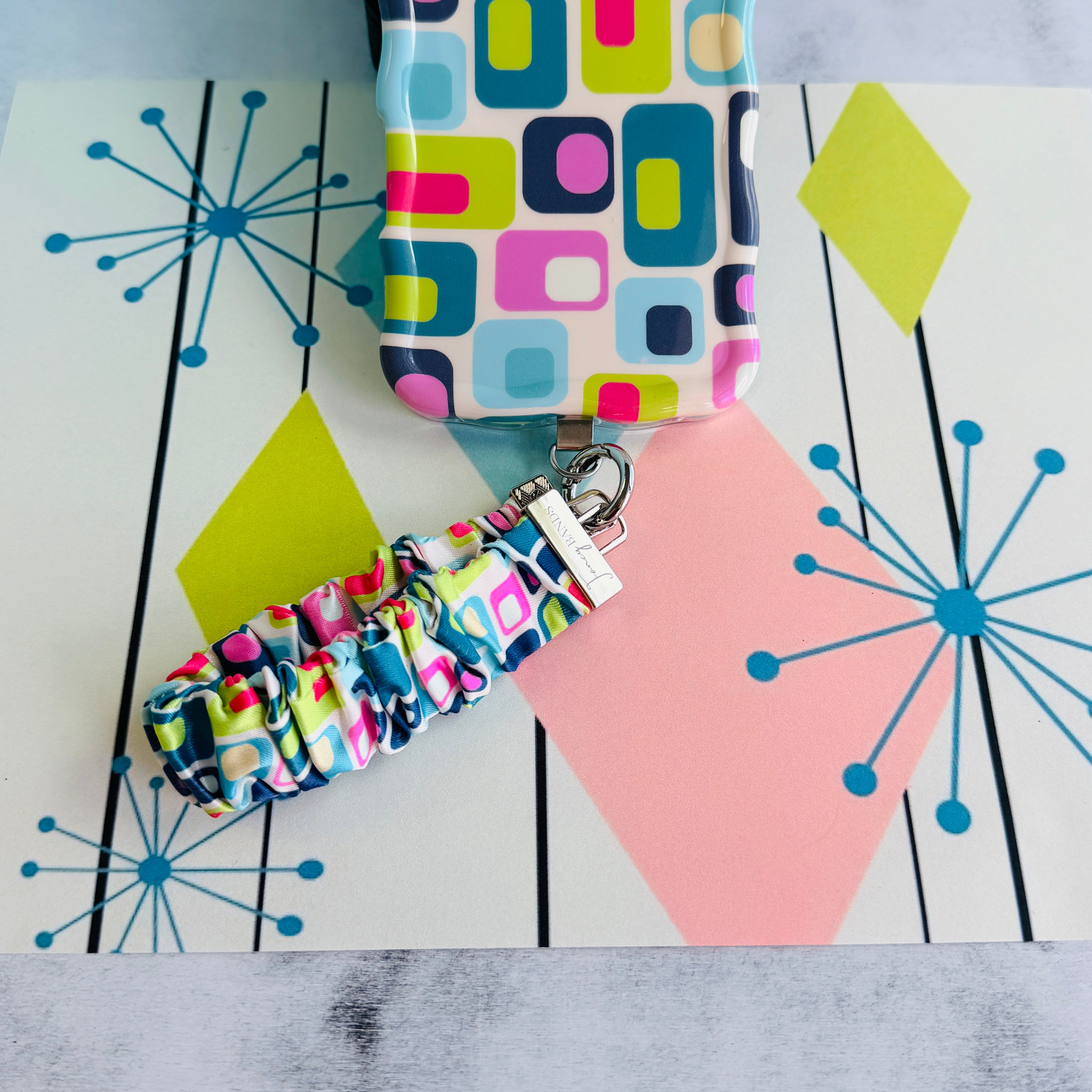 Mod Mood Scrunchie Phone Wrist Strap clipped to the matching retro geometric phone case, on an atomic starburst backdrop.