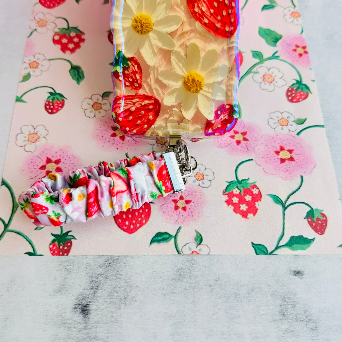 Berry Cute Blooms scrunchie phone wrist strap and matching strawberry and daisy Wavy Luxe iPhone case laid out together on strawberry print paper