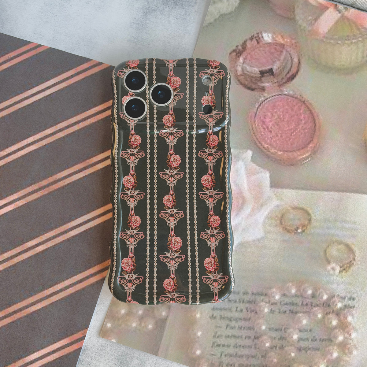 The Pretty Little Details MagSafe iPhone case laid on black and rose gold striped paper beside a pink blush compact and a strand of pearls.