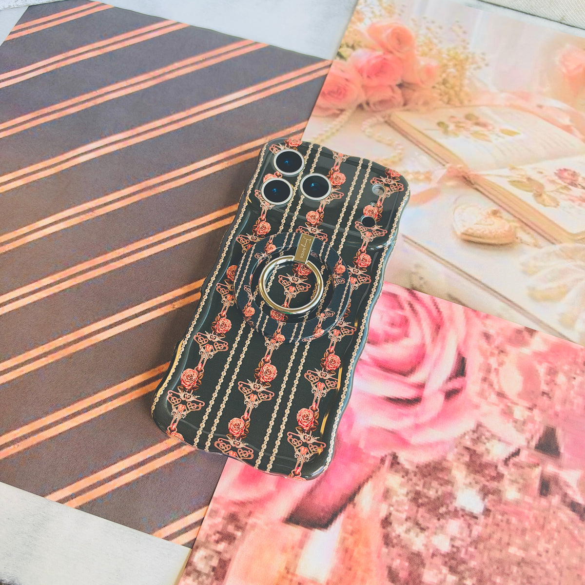 Pretty Little Details black pearl and rose iPhone case wearing the matching MagSafe grip ring, styled on striped paper with pink rose prints.