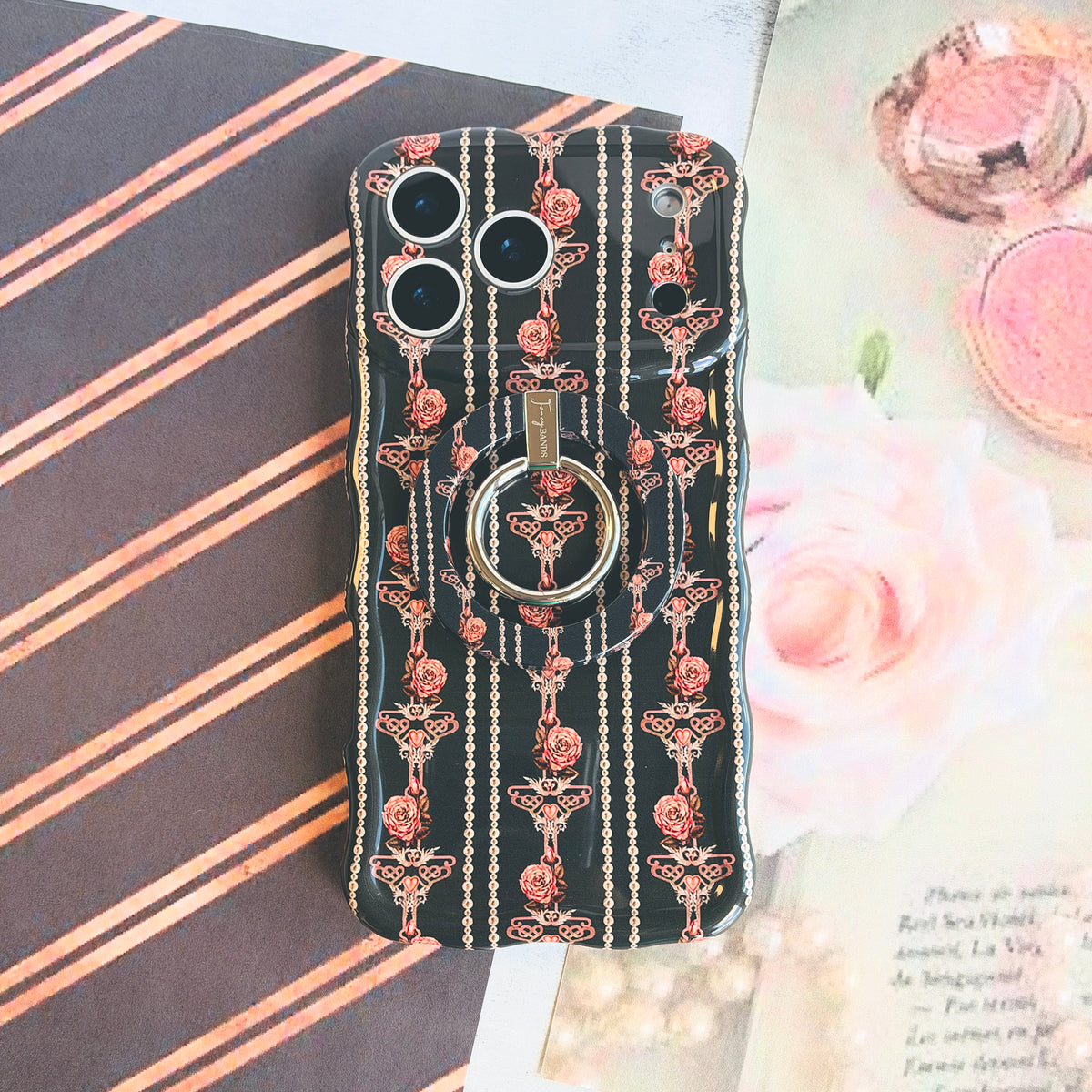 The Pretty Little Details black pearl and rose MagSafe grip ring attached to the matching iPhone case, with a pink compact and pearls on a striped flat lay.