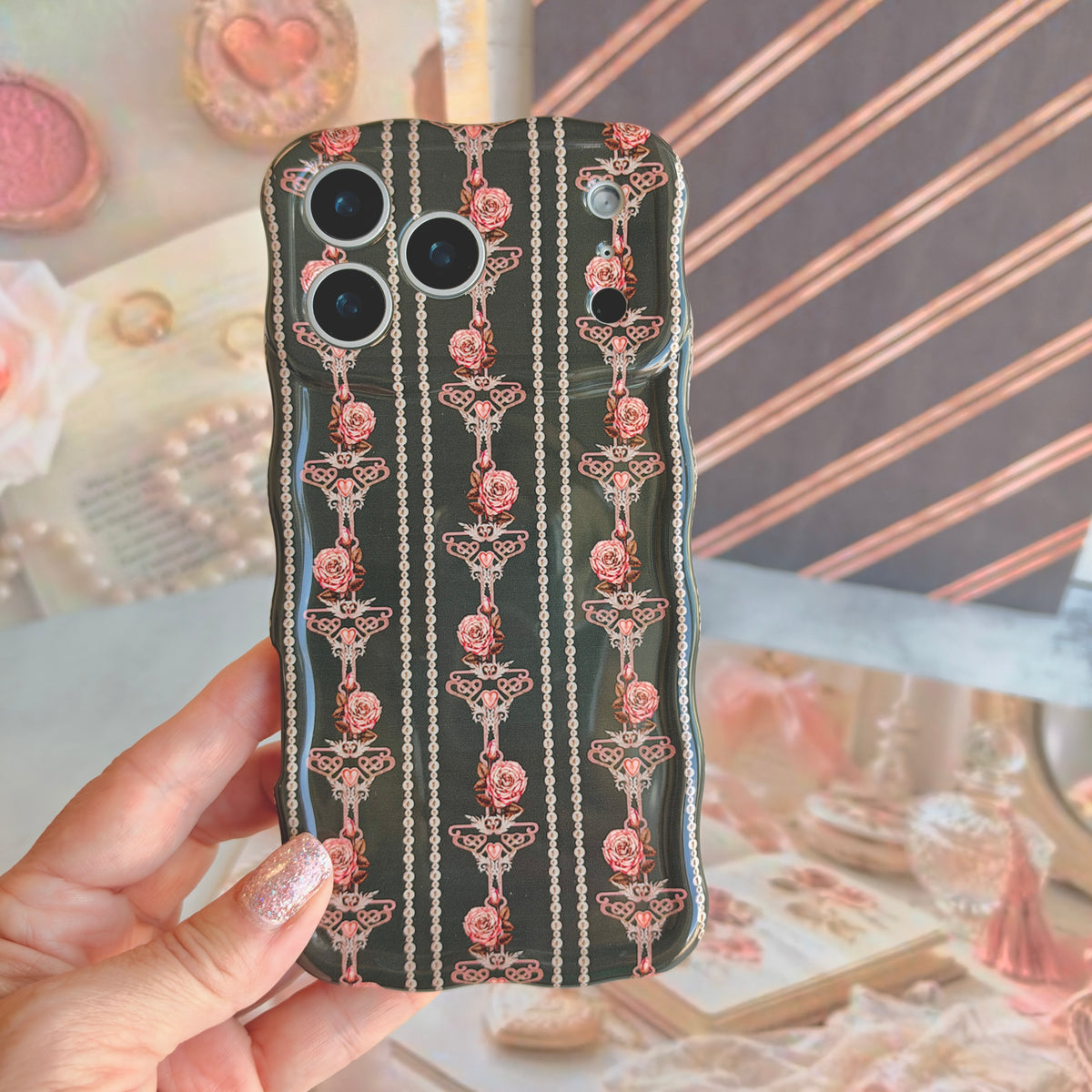 Hand holding the Pretty Little Details black pearl and rose Wavy Luxe iPhone case over a coquette flat lay with a blush compact, pearls and pink roses.