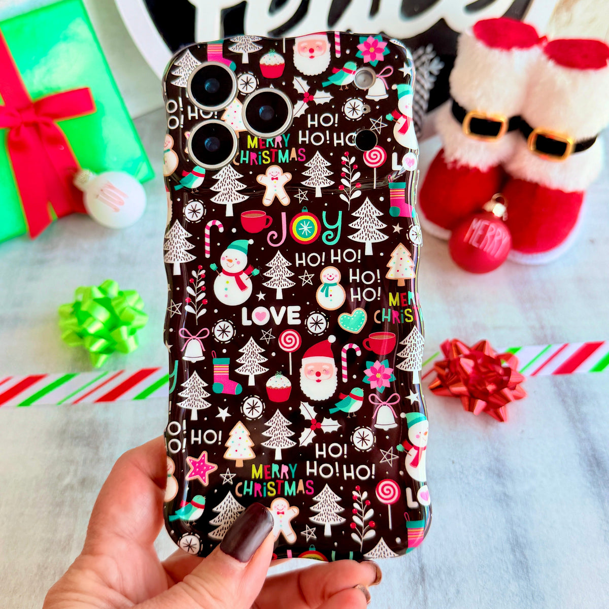 Hand holding the Jolly Wrist-mas Wavy Luxe MagSafe iPhone case, Santas, snowmen and HO HO HO lettering on black