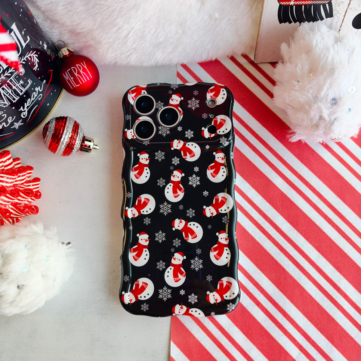 Snowman Wavy Luxe MagSafe iPhone case laid flat, black snowman print, on candy cane stripes with a Santa hat and ornaments