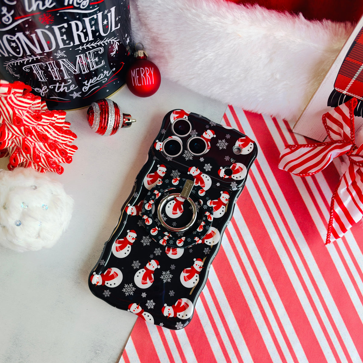 Snowman iPhone case with the matching MagSafe grip ring attached, on candy cane stripes with a Santa hat and ornaments