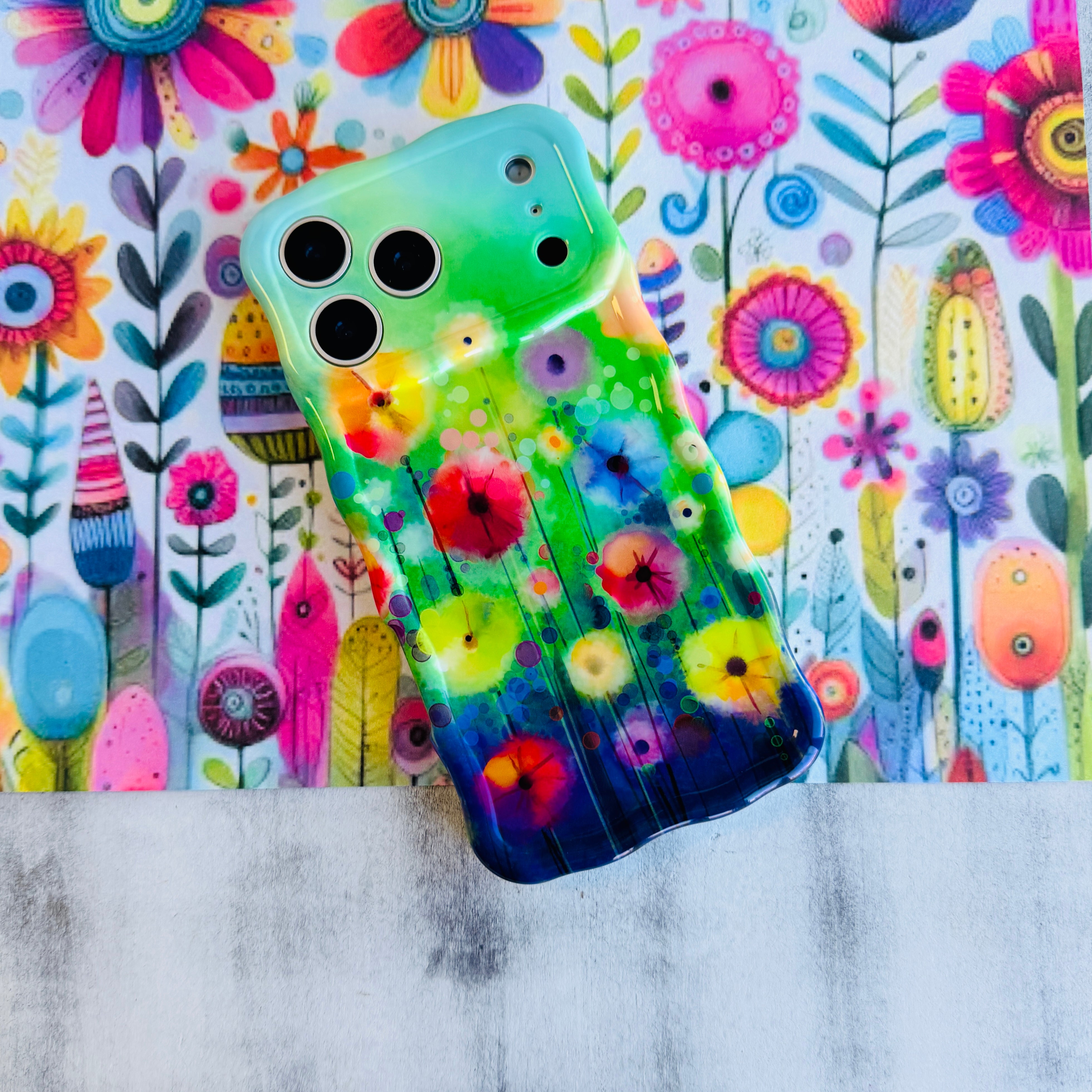 Wavy iPhone case featuring vibrant watercolor flowers in pink, red, yellow, and orange on a green and blue gradient background.