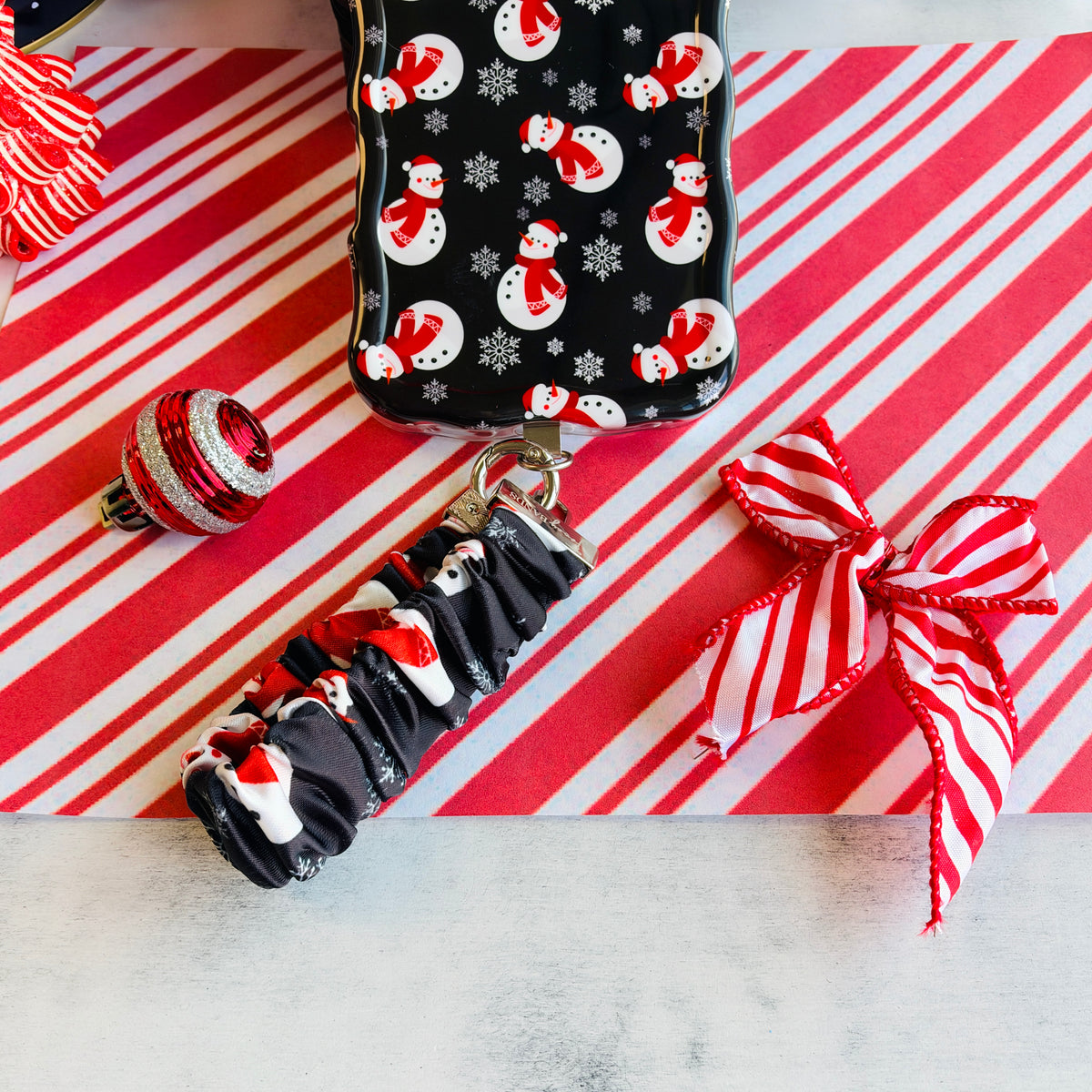 Snowman scrunchie phone wrist strap clipped to the matching iPhone case, on candy cane stripes with a Santa hat and ornaments