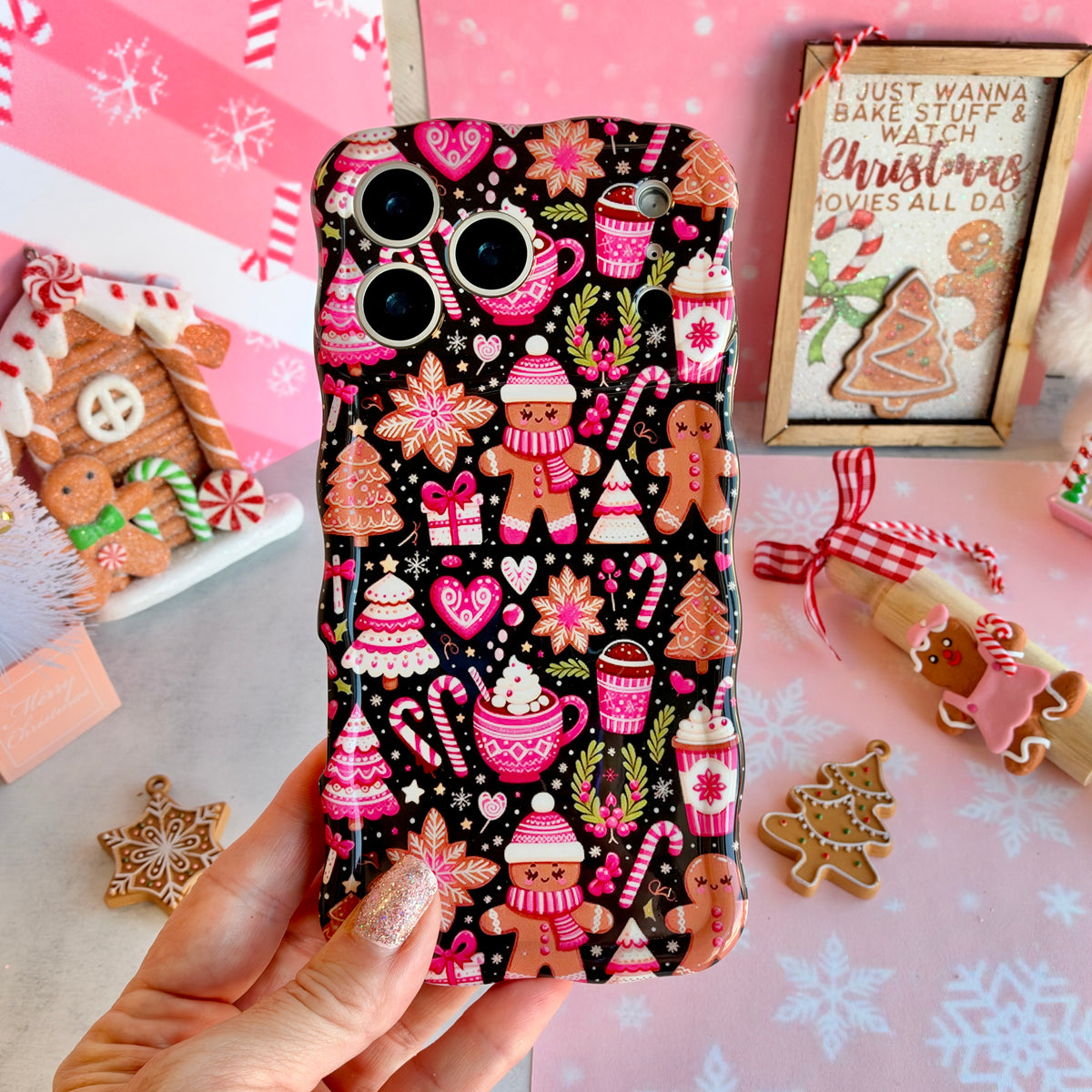 Hand holding the Merry Mugs & Ginger Hugs Wavy Luxe MagSafe iPhone case in pink gingerbread and cocoa mug print on black, with gingerbread cookies and a gingerbread house