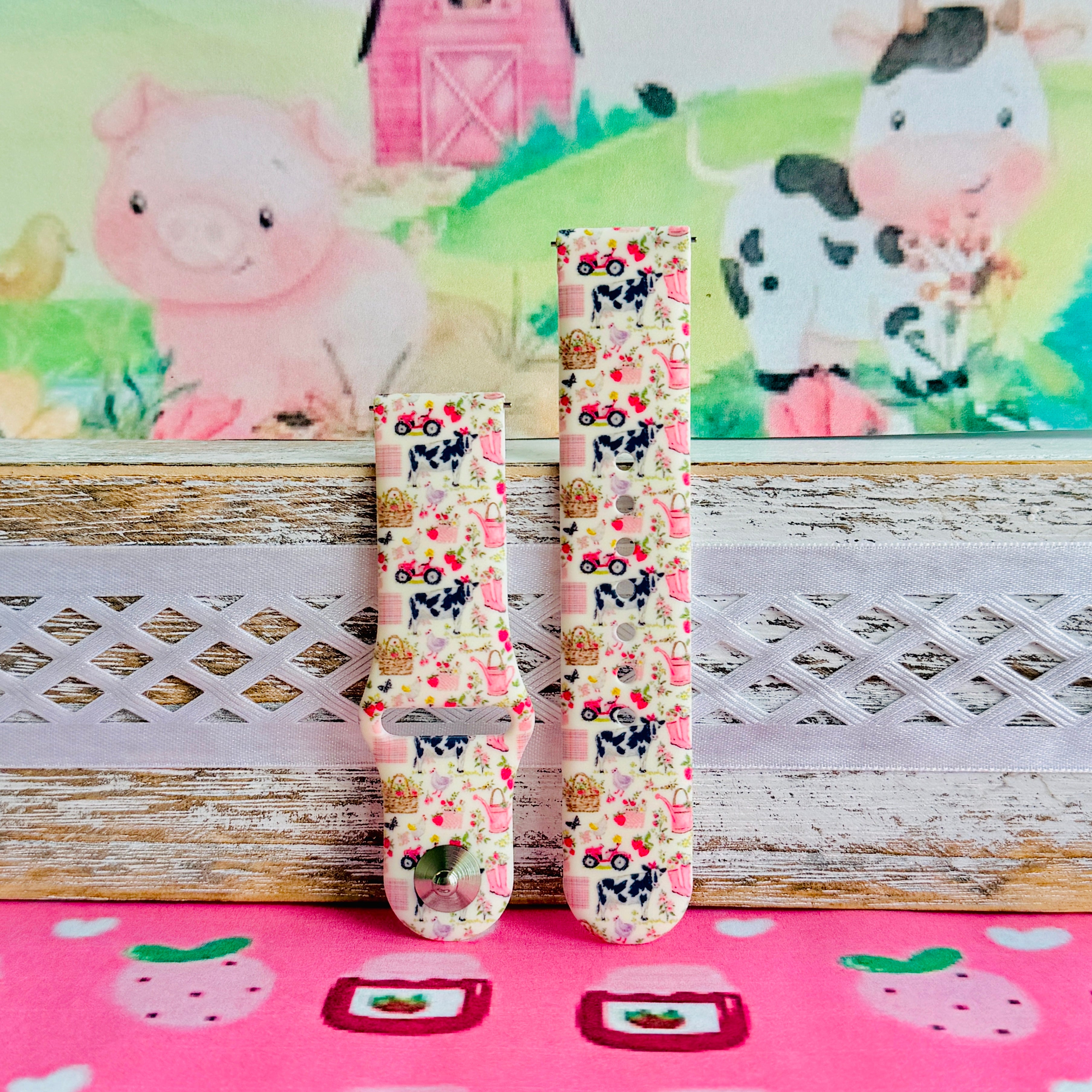 Farm Girl Cutie silicone Samsung Galaxy Watch band with dairy cows, red tractors, chickens, ducks, pink watering cans, strawberries, baskets, and roses on a cream base with pink gingham squares, shown displayed and worn against a farmhouse country backdrop.