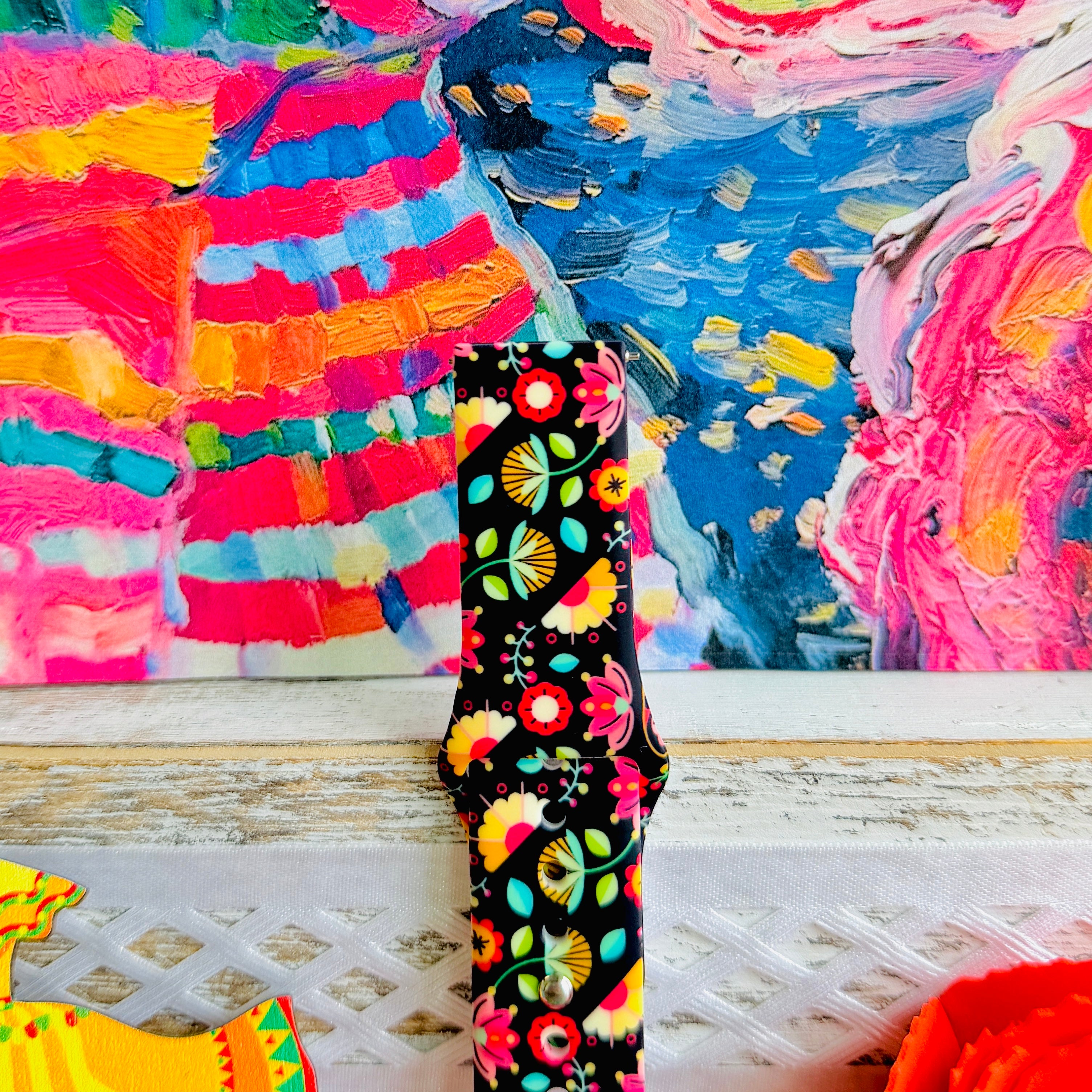 Bloom Bonita silicone Samsung Galaxy Watch band with Mexican folk art florals in coral, red, yellow, teal, and pink on a black base, shown displayed against a vibrant Mexican folk art backdrop with rose and dress accents.