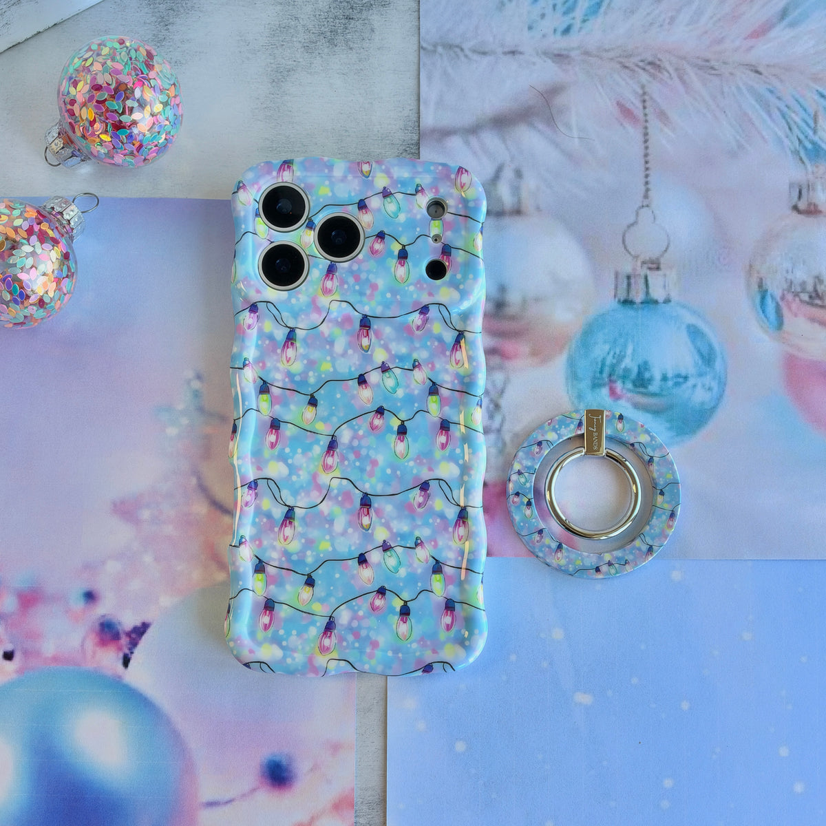 Twinkle All The Way MagSafe grip ring lying beside the matching Christmas lights iPhone case and a blue glass ornament