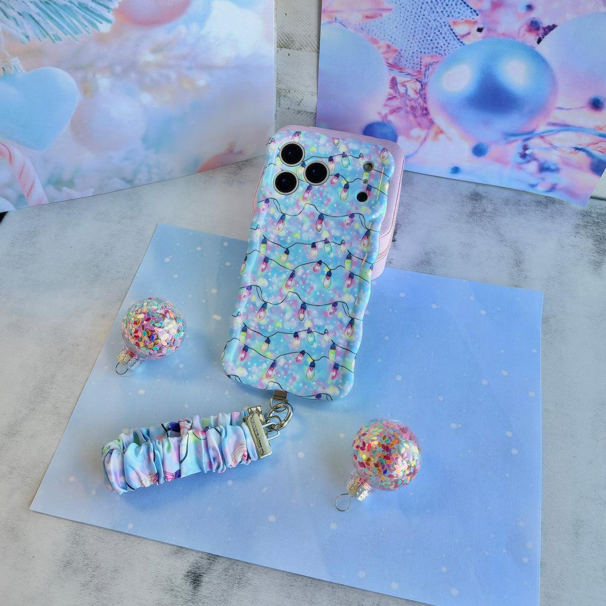 Angled flat lay of the Twinkle All The Way iPhone case with its Christmas lights scrunchie wrist strap on pastel blue paper