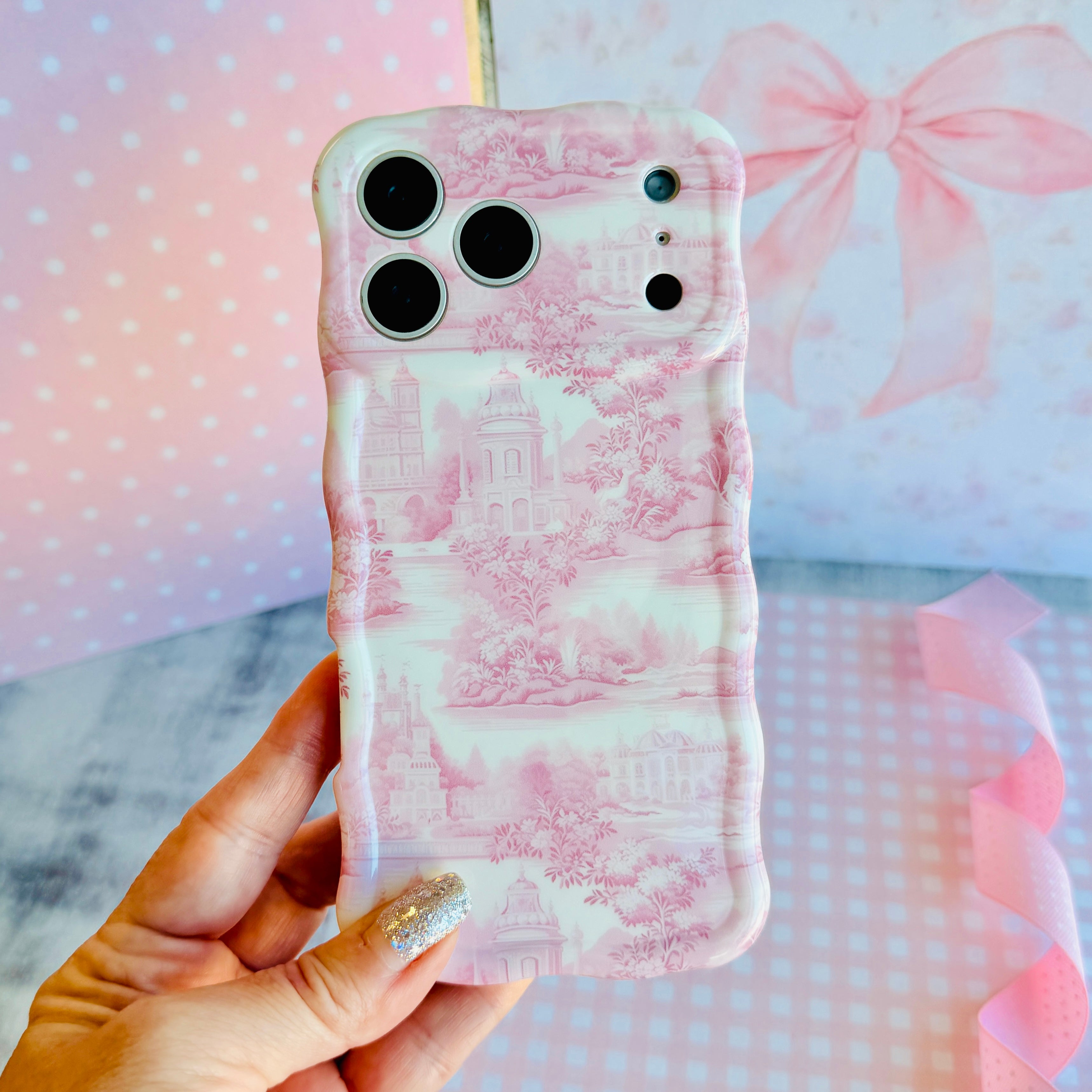 Hand holding the Blush Manor Wavy Luxe MagSafe iPhone case, pink toile manor houses and gardens on a cream ground, against a pink bow backdrop