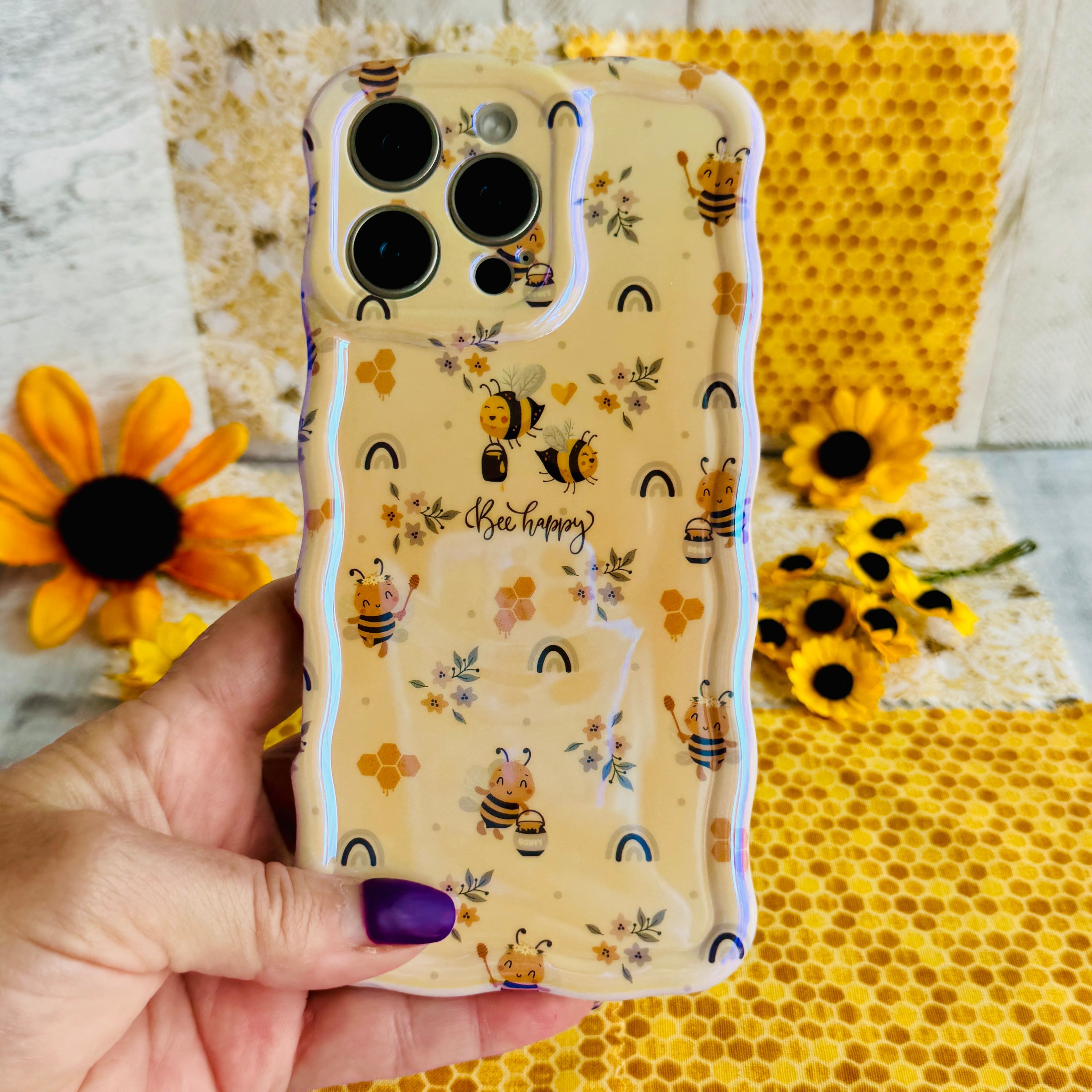 Hand holding the Bee Happy Wavy Luxe MagSafe iPhone case with honeybees and rainbows on a honey-cream background
