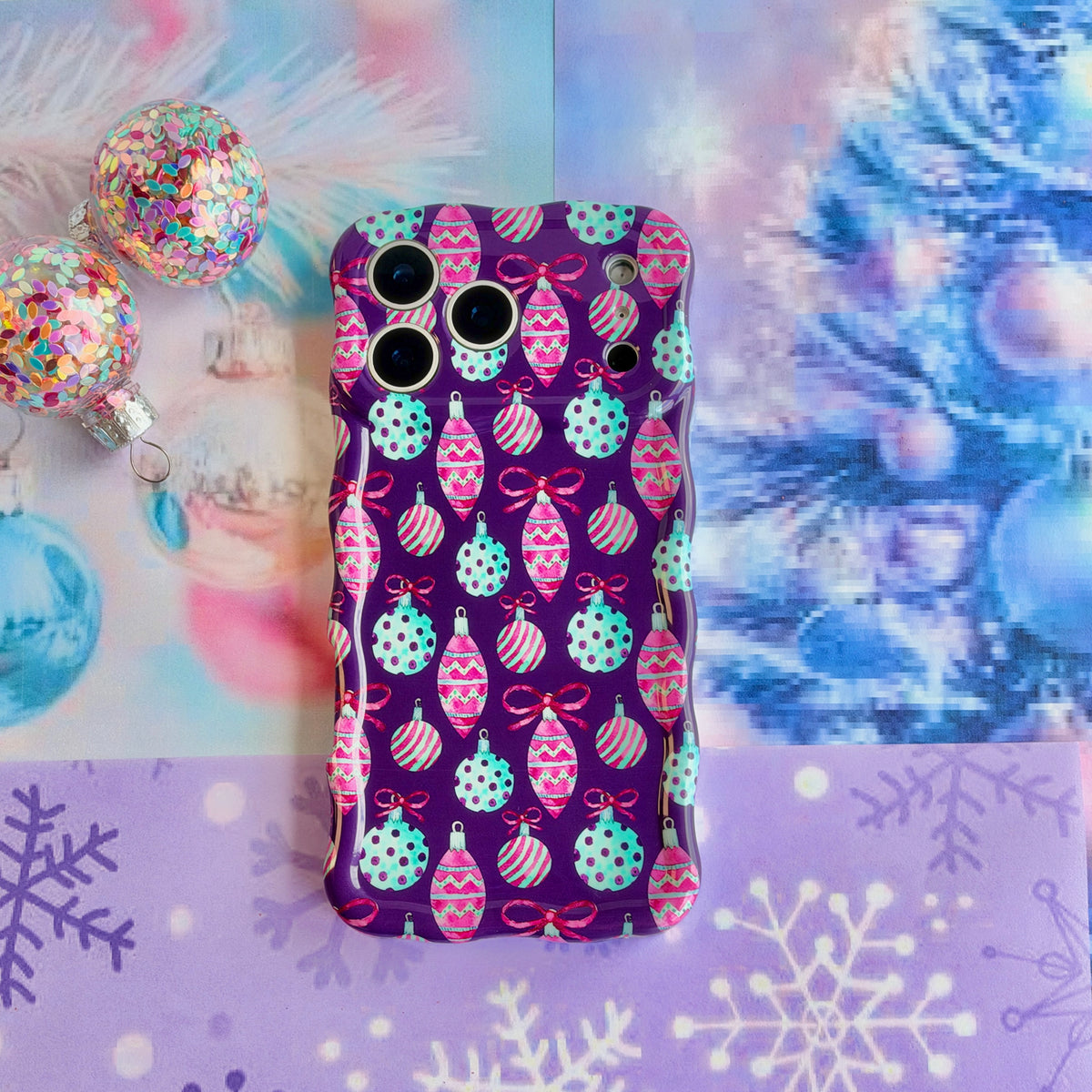 Just One More Ornament iPhone case lying flat on lavender snowflake paper beside aqua tinsel and a confetti-filled clear ornament