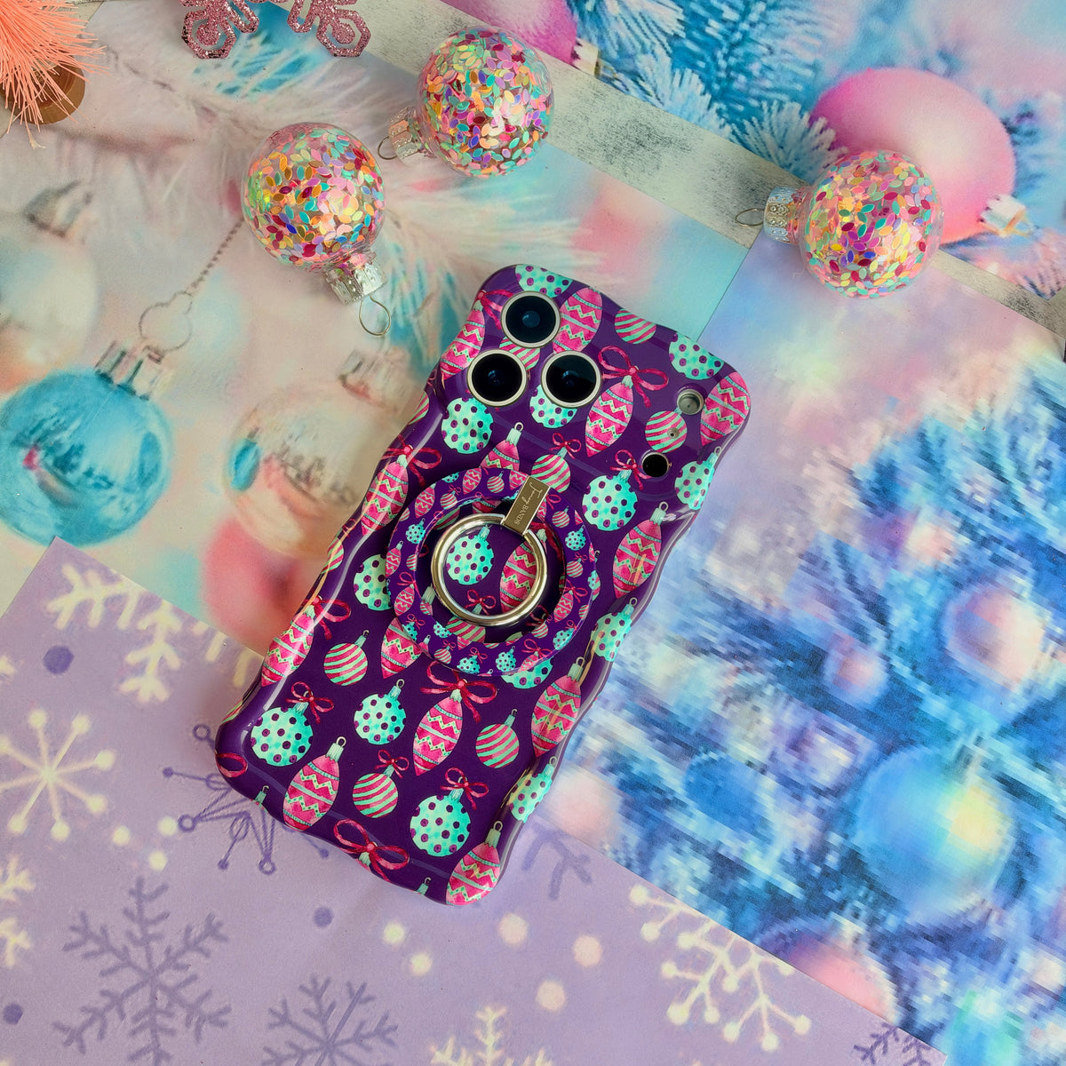 Just One More Ornament iPhone case with the matching grip ring snapped on, resting on aqua tinsel with pink glitter snowflakes and glass baubles