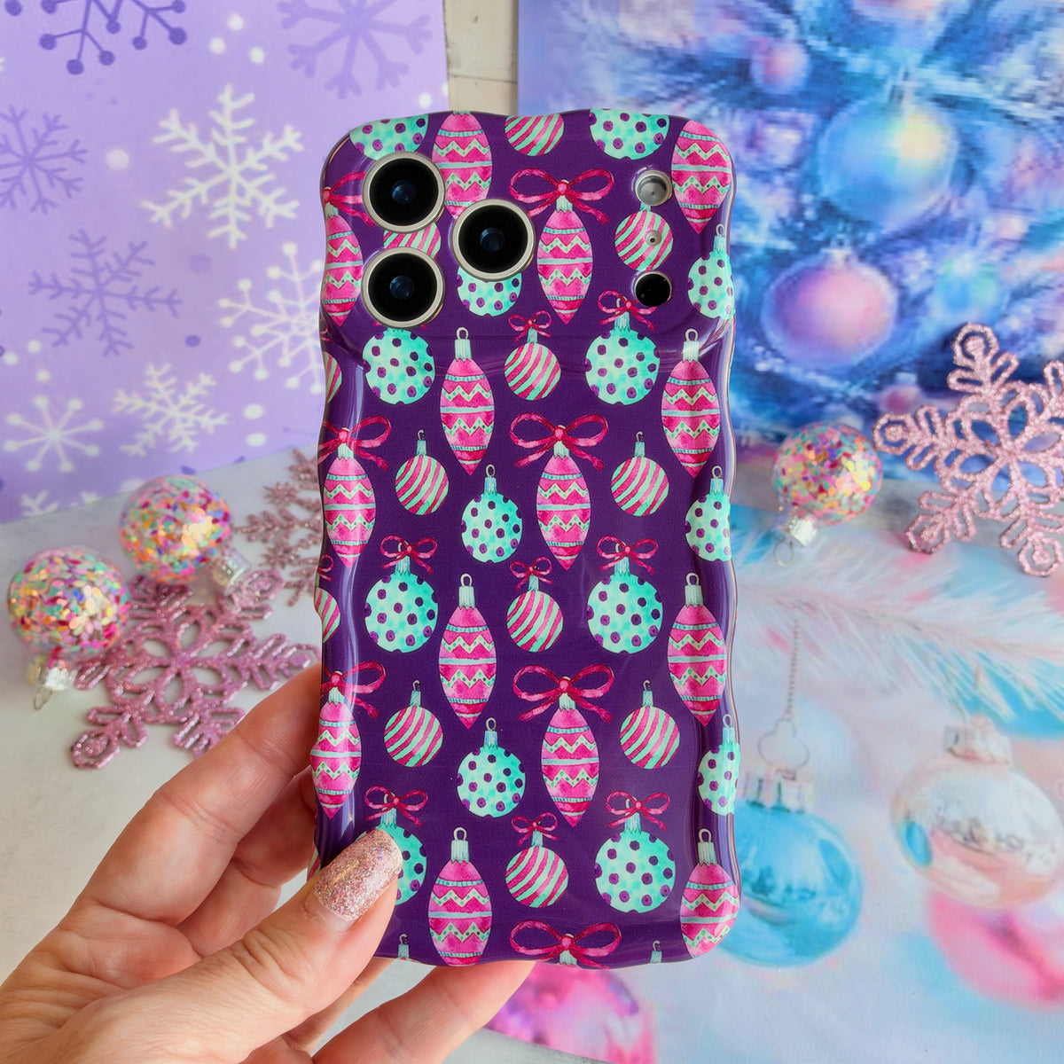 Hand holding the Just One More Ornament Wavy Luxe MagSafe iPhone case, pink and aqua watercolor Christmas ornaments on plum, in front of lavender snowflakes and an aqua tinsel tree
