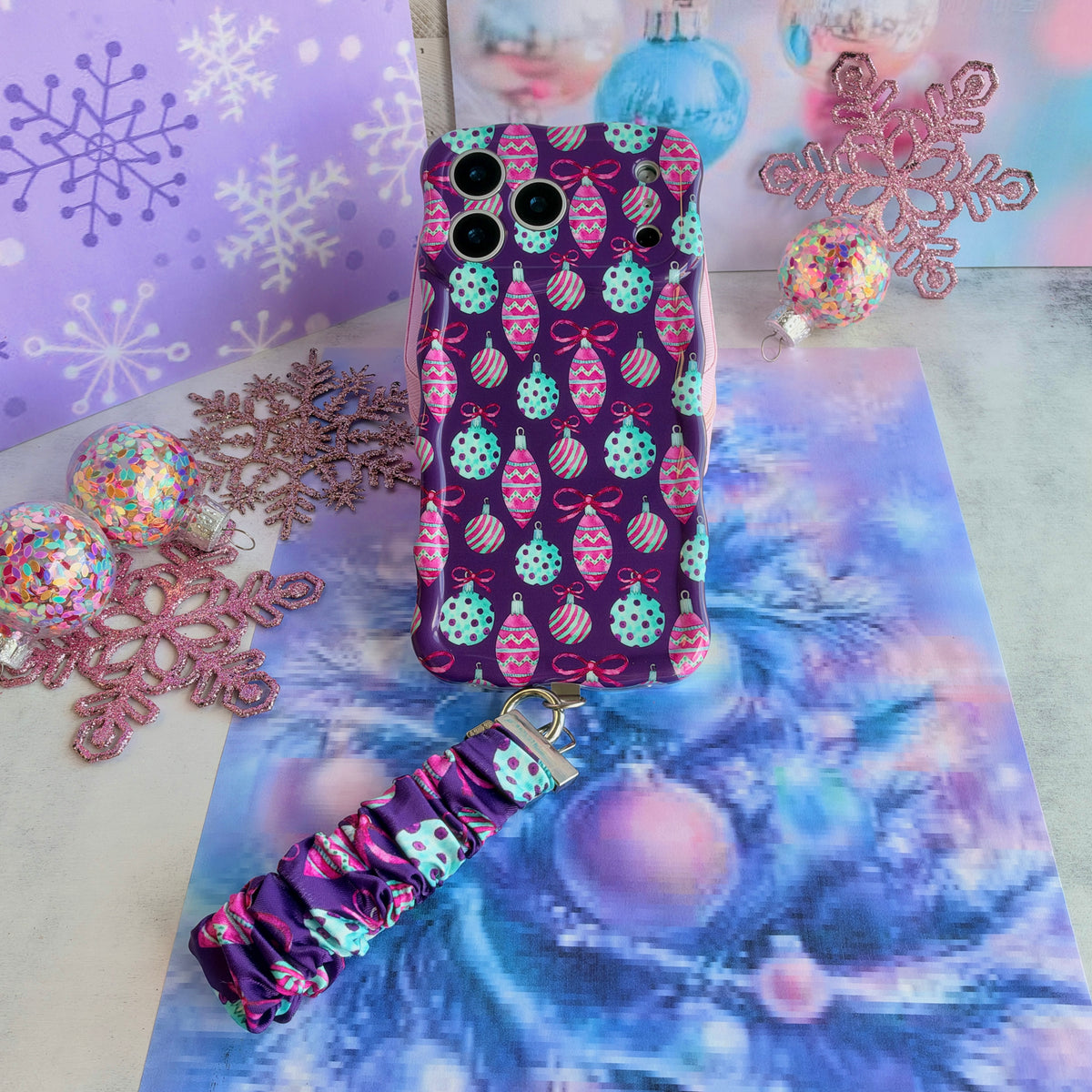 Just One More Ornament iPhone case with its ornament-print scrunchie wrist strap on lavender snowflake paper and aqua tinsel, beside a pink glitter snowflake