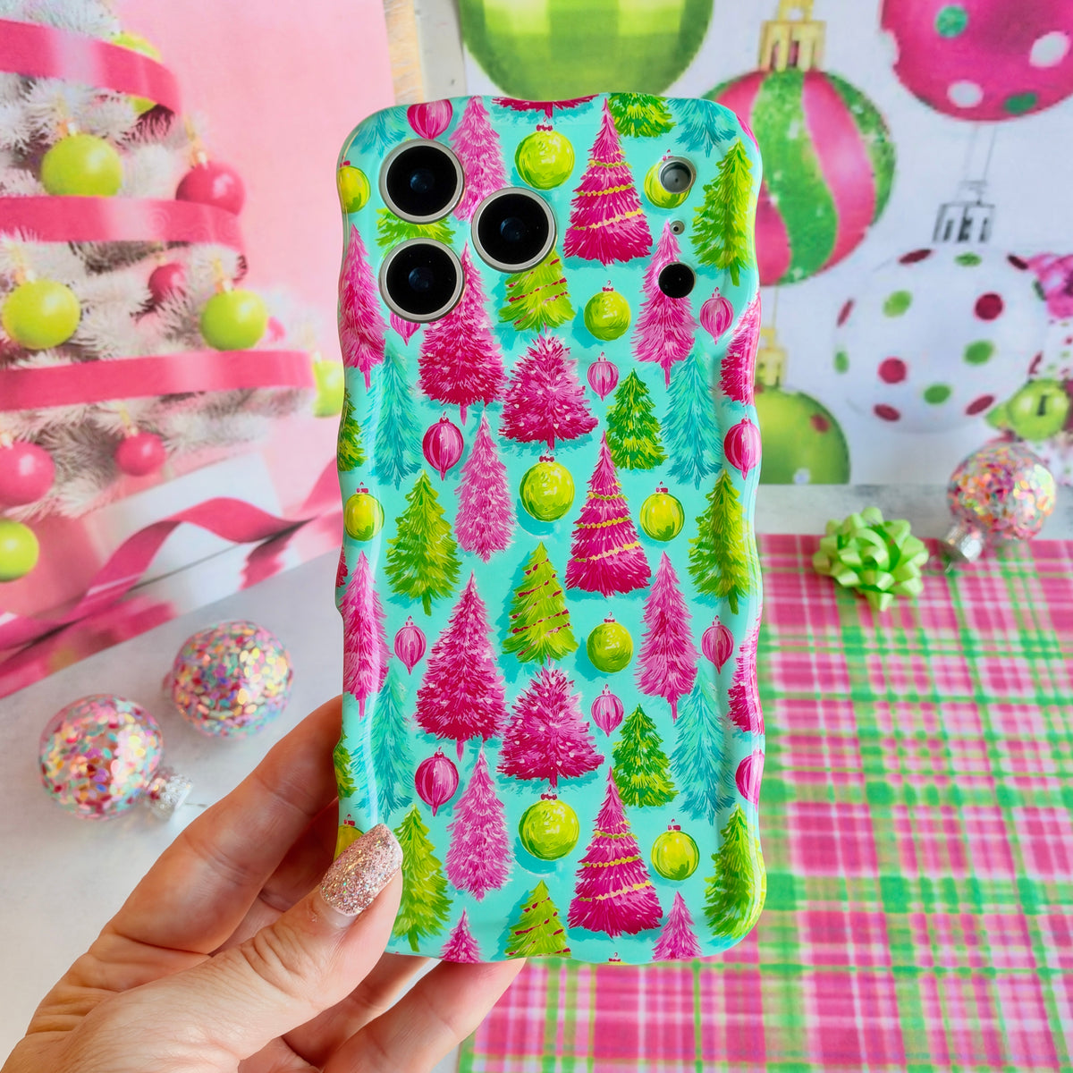 Hand holding the Merry & Bright, Totally! Wavy Luxe MagSafe iPhone case, hot pink and lime bottle-brush Christmas trees on aqua, in front of big pink and green ornaments