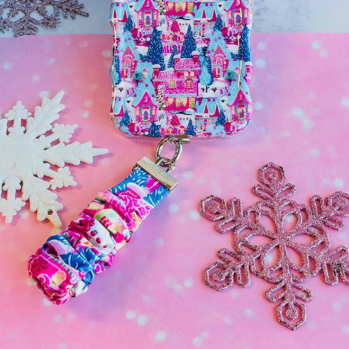 Sugarplum Square scrunchie phone wrist strap clipped below the matching iPhone case on pink paper with white and pink glitter snowflakes