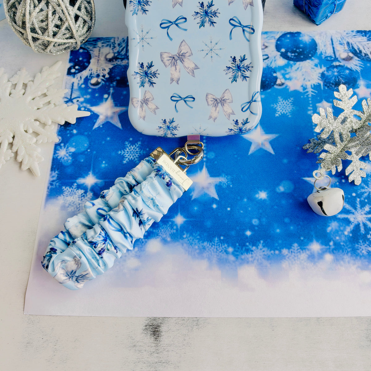 Snow Pretty In Blue scrunchie phone wrist strap clipped below the matching iPhone case on blue snowflake paper with glitter snowflakes