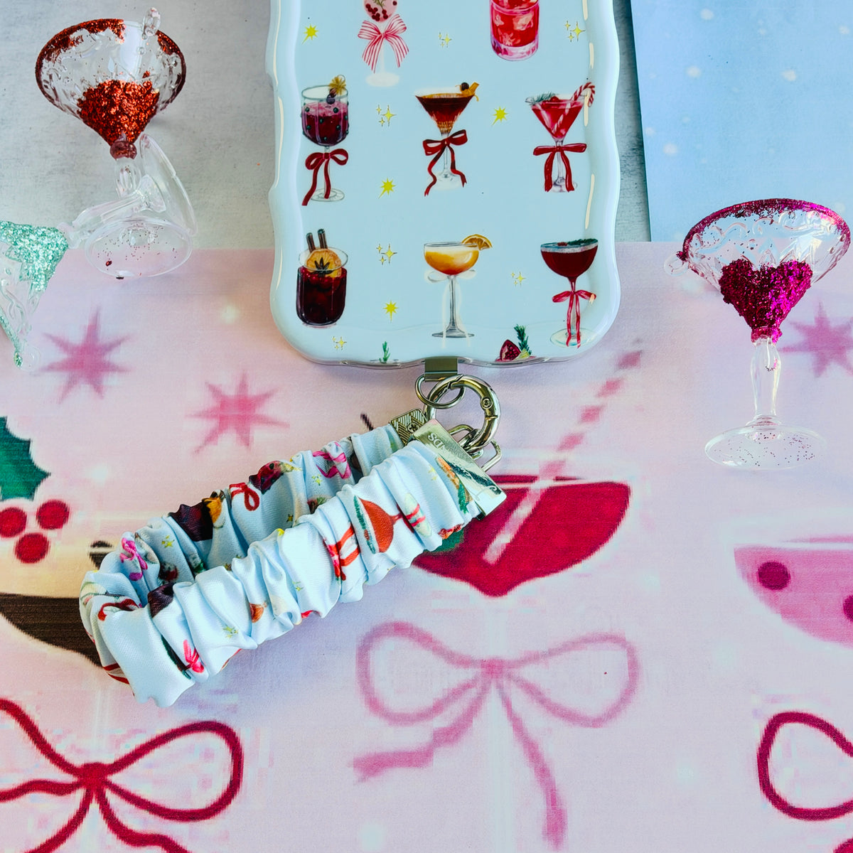 Holiday Spirits, Please? scrunchie phone wrist strap clipped below the matching iPhone case on pink cocktail paper with a glitter-rimmed glass ornament