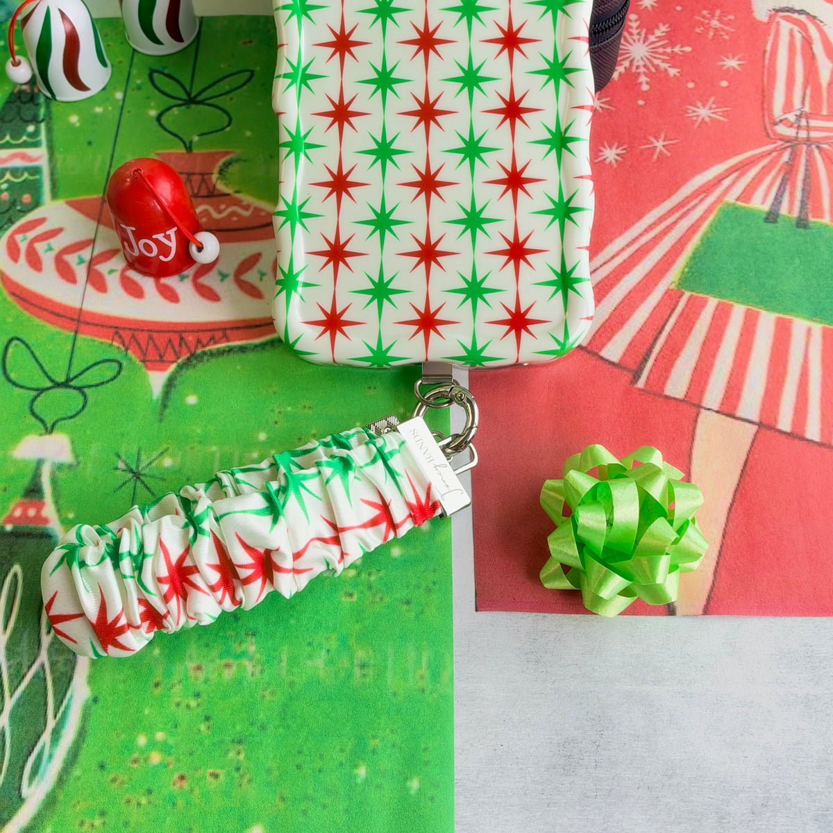 Tinsel & Stardust scrunchie phone wrist strap clipped below the matching starburst iPhone case, with a Joy ornament and green gift bow
