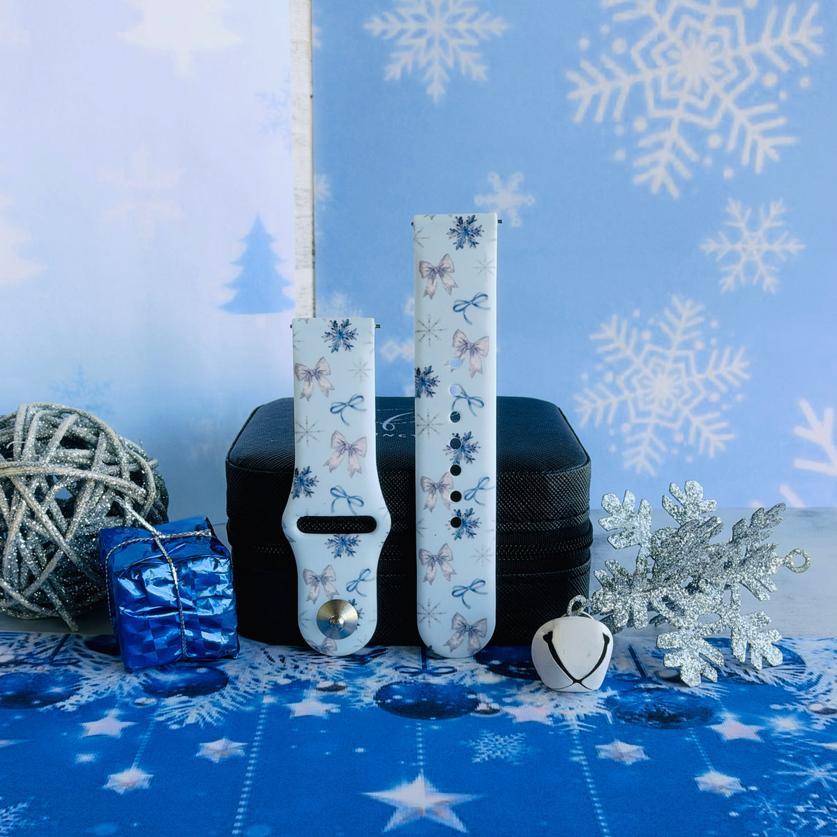 Both straps of the Snow Pretty In Blue Samsung Galaxy Watch band on a jewelry box with a white jingle bell on a blue starry snowflake backdrop