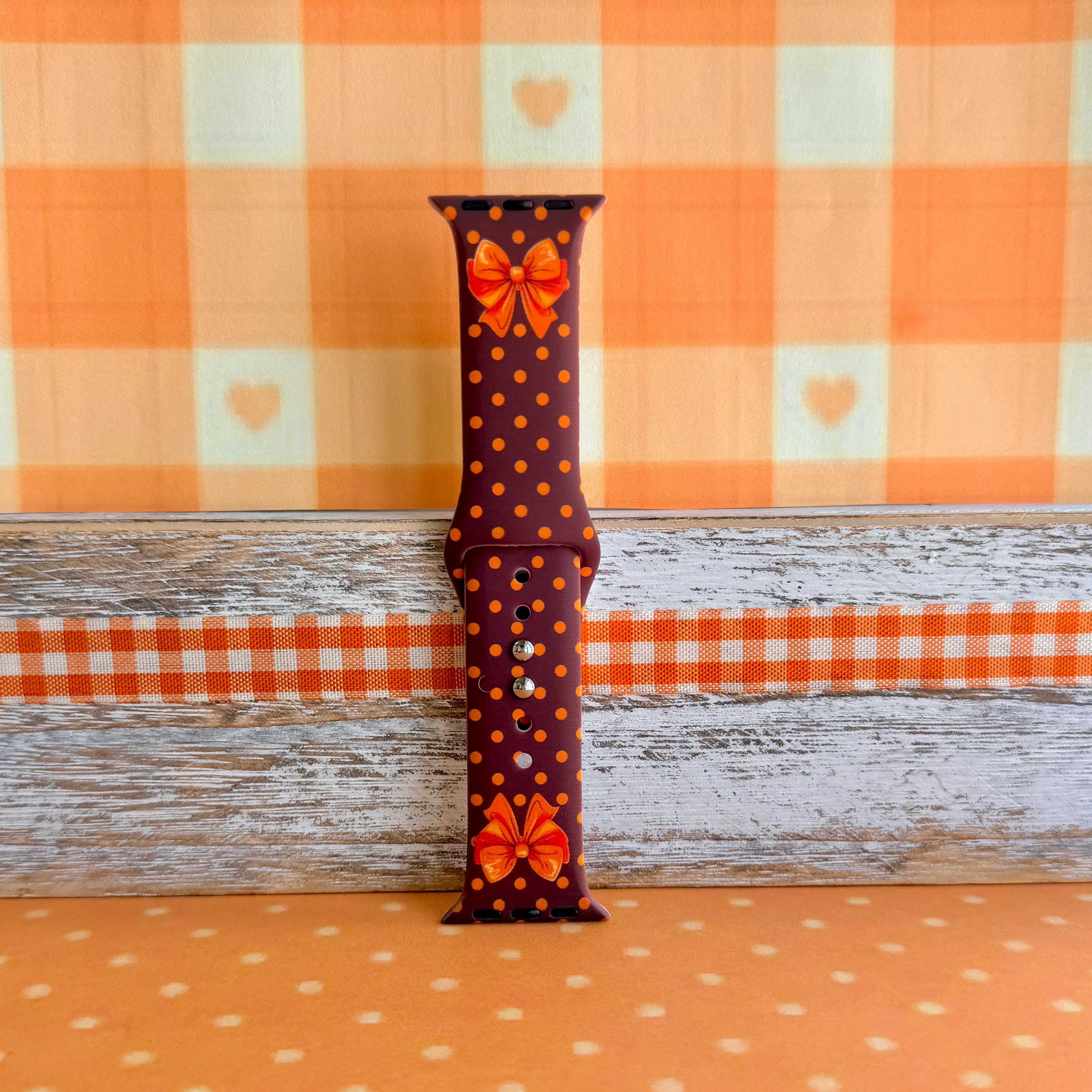 Spotted Looking Cute Apple Watch band single strap half standing against orange gingham, showing the orange bow detail and pumpkin dots on chocolate brown silicone.
